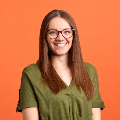 A young woman with glasses, long brown hair, and a green shirt smiling against an orange background.