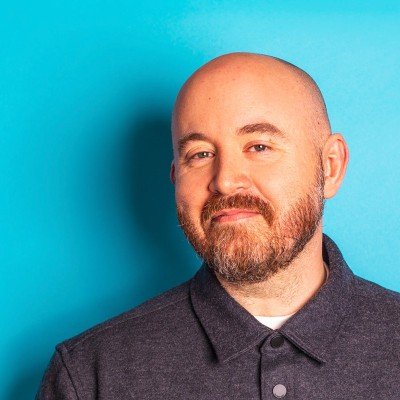 A man with a beard and bald head, wearing a dark collared shirt, standing against a bright blue background.