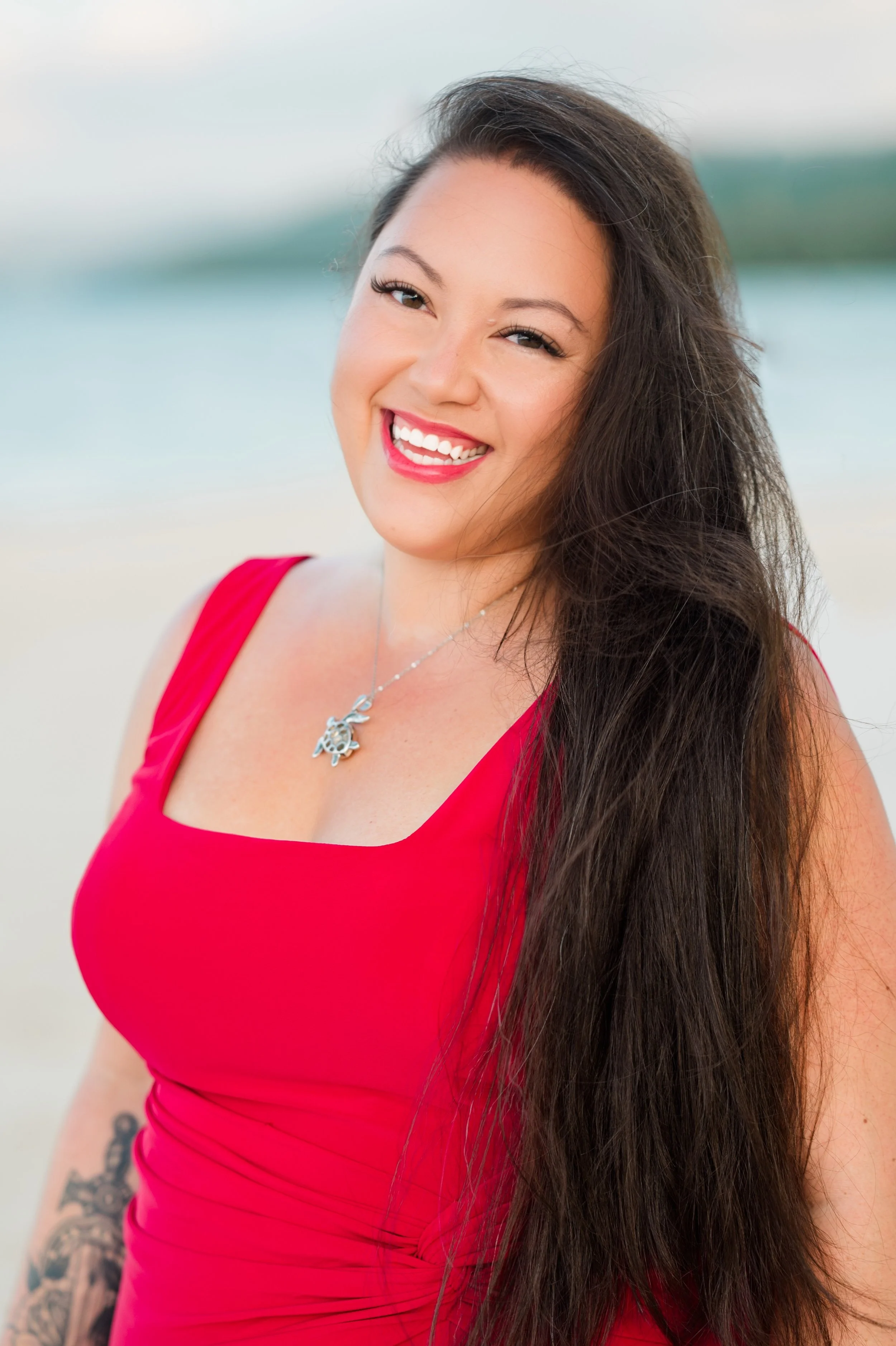 A smiling woman with dark, long hair, wearing a red sleeveless dress, standing outdoors with a blurred water and sky background.