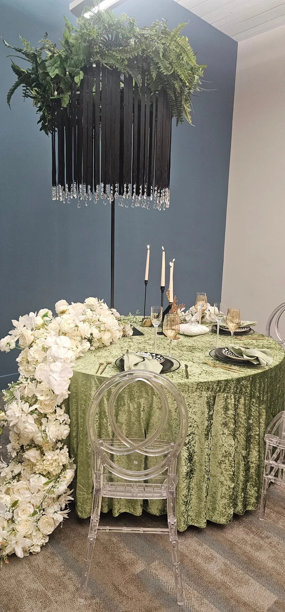A decorated dining table with a green textured tablecloth, white floral centerpiece, black plates, and clear glassware. Tall white candles in candlesticks and an inverted candle holder are on the table. A black chandelier with green plants and hanging crystals hangs above, and transparent chairs surround the table.