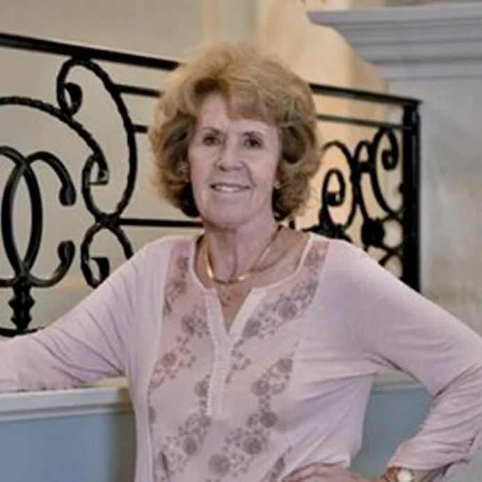 A woman with short, curly blonde hair wearing a light pink blouse with embroidery, standing indoors with an ornate black wrought iron railing behind her.