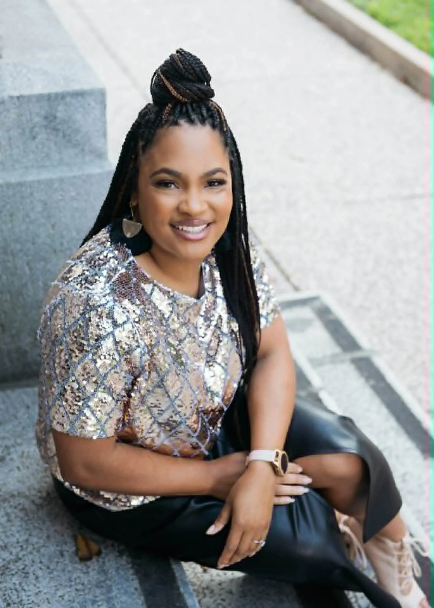 A woman sitting on outdoor steps, smiling, with braided hair styled in a top bun, wearing a patterned top, black pants, and light-colored sneakers.