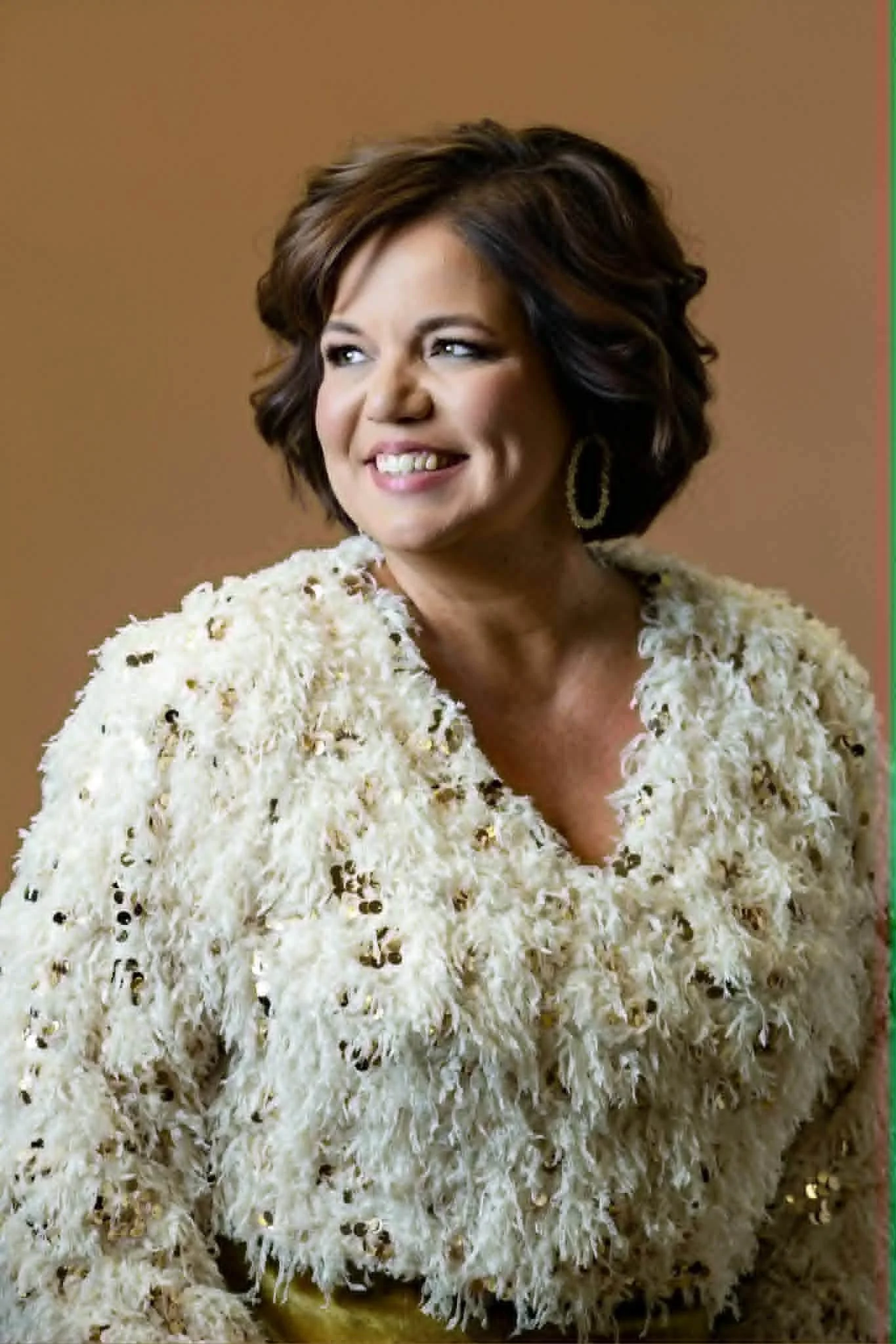 A woman with short, wavy brown hair with highlights, smiling and wearing a cream-colored textured coat with black accents and gold earrings.