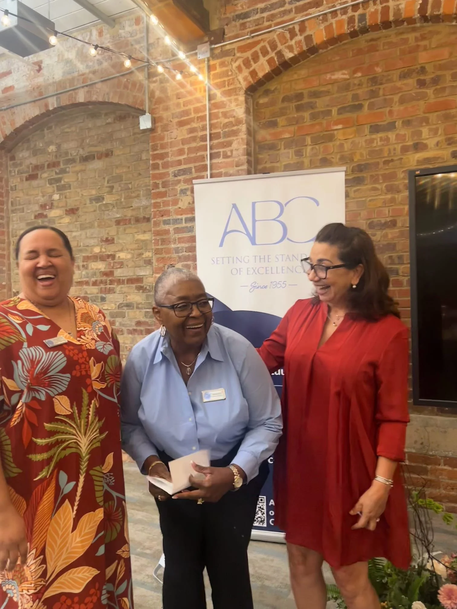 Three women standing together at an event, smiling and laughing. Behind them is a banner with the letters 'ABC' and the phrase 'Setting the Standard of Excellence Since 1955'. The background features brick walls and string lights.