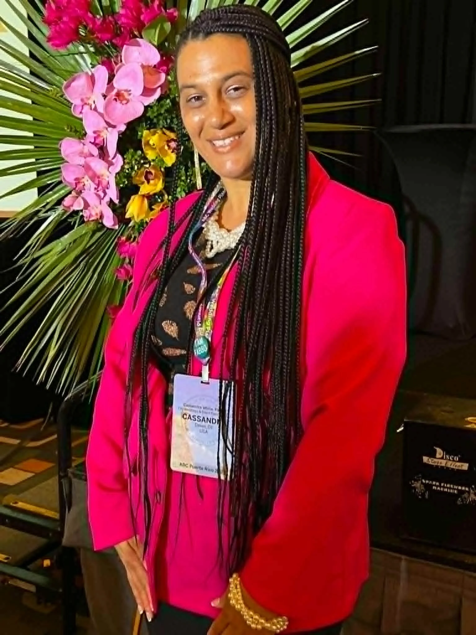 A woman with long braided hair standing in front of a background of pink and yellow orchids and large green tropical leaves. She is wearing a bright pink blazer, a black top, a pearl necklace, and a name badge hanging from a lanyard.
