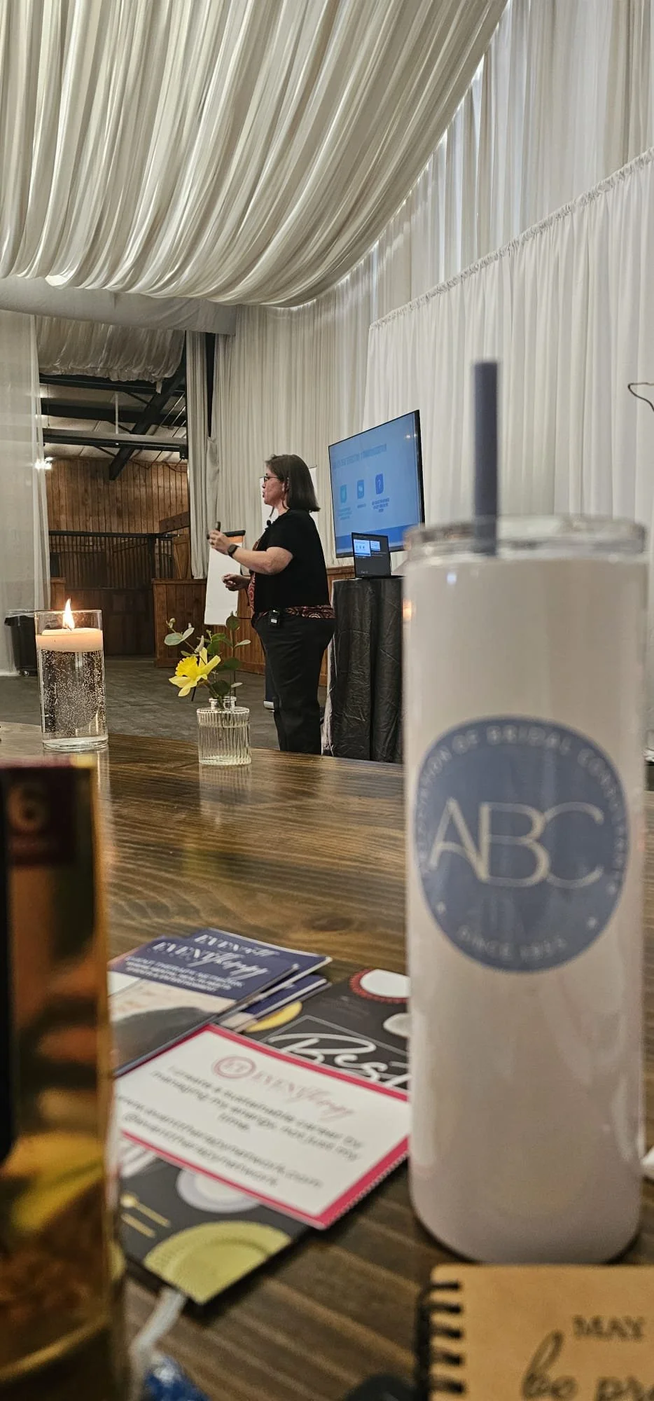 A woman giving a presentation in a room with white draped ceiling and walls, a large screen television, a table with pamphlets and drinks, and a candle in the foreground.
