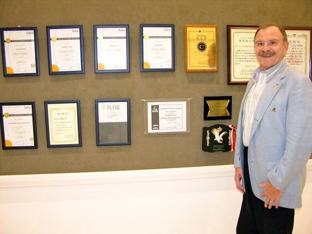 A man in a light blue blazer and white shirt standing in front of a wall with multiple certificates and awards displayed. The certificates are framed and arranged in two rows, with one award in a gold frame and another in a black frame. The man is smiling and has short hair and a mustache.
