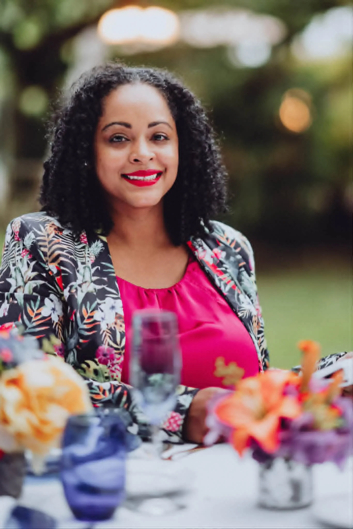 A woman with curly black hair and red lipstick smiling at a gathering, wearing a pink top and a floral blazer, with blurred flowers and tableware in front of her.