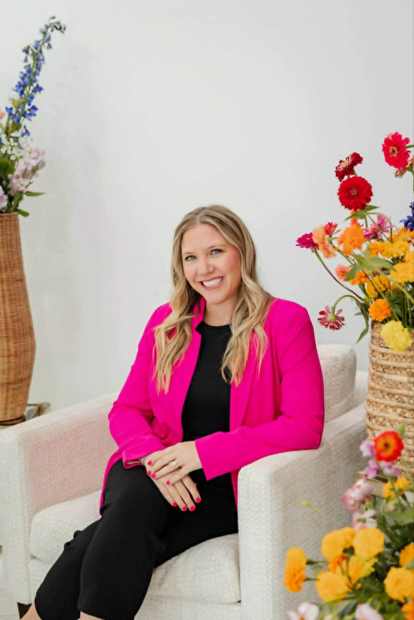 A smiling woman with long blonde hair, wearing a bright pink blazer and black outfit, sitting on a white upholstered chair, surrounded by colorful flowers in vases.
