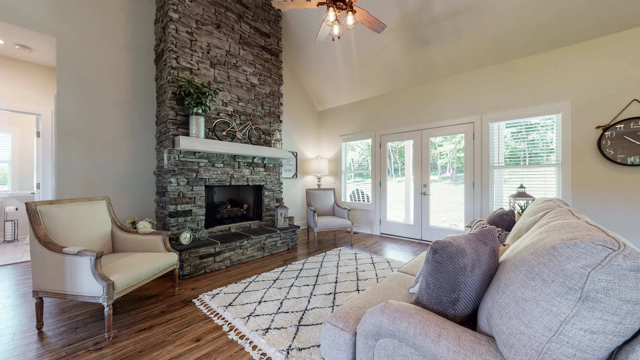 Living room featuring a stone fireplace, beige sofa, two armchairs, decorative throw pillows, wooden floor, and large windows with a view of the outdoors.