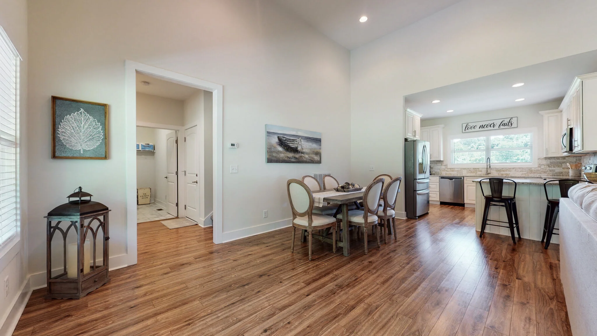 Open-concept dining and kitchen area with a wooden table, six chairs, hardwood floor, framed art on the wall, modern kitchen with stainless steel appliances, and a sign reading 'love never fails.'
