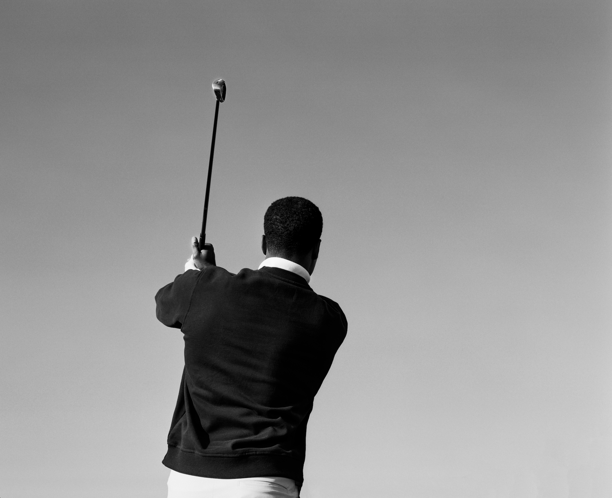 A man dressed in a dark sweater with a white collar shirt holding a golf club and preparing to hit a golf ball against a cloudy sky.