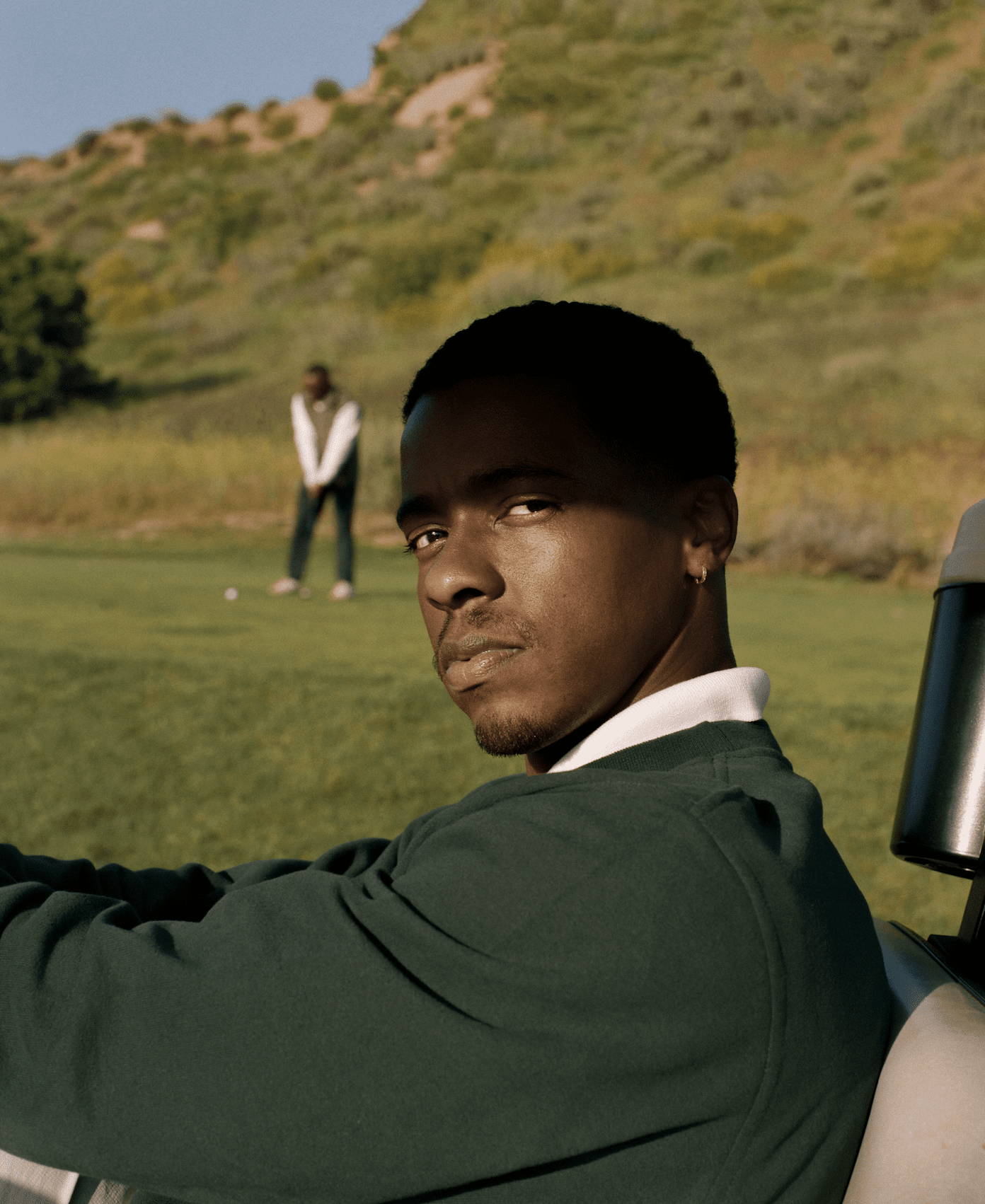 A young man with dark skin and short black hair sitting in a golf cart on a golf course, looking over his shoulder with a serious expression, while another man in a white shirt and vest stands in the background preparing to take a shot. The landscape