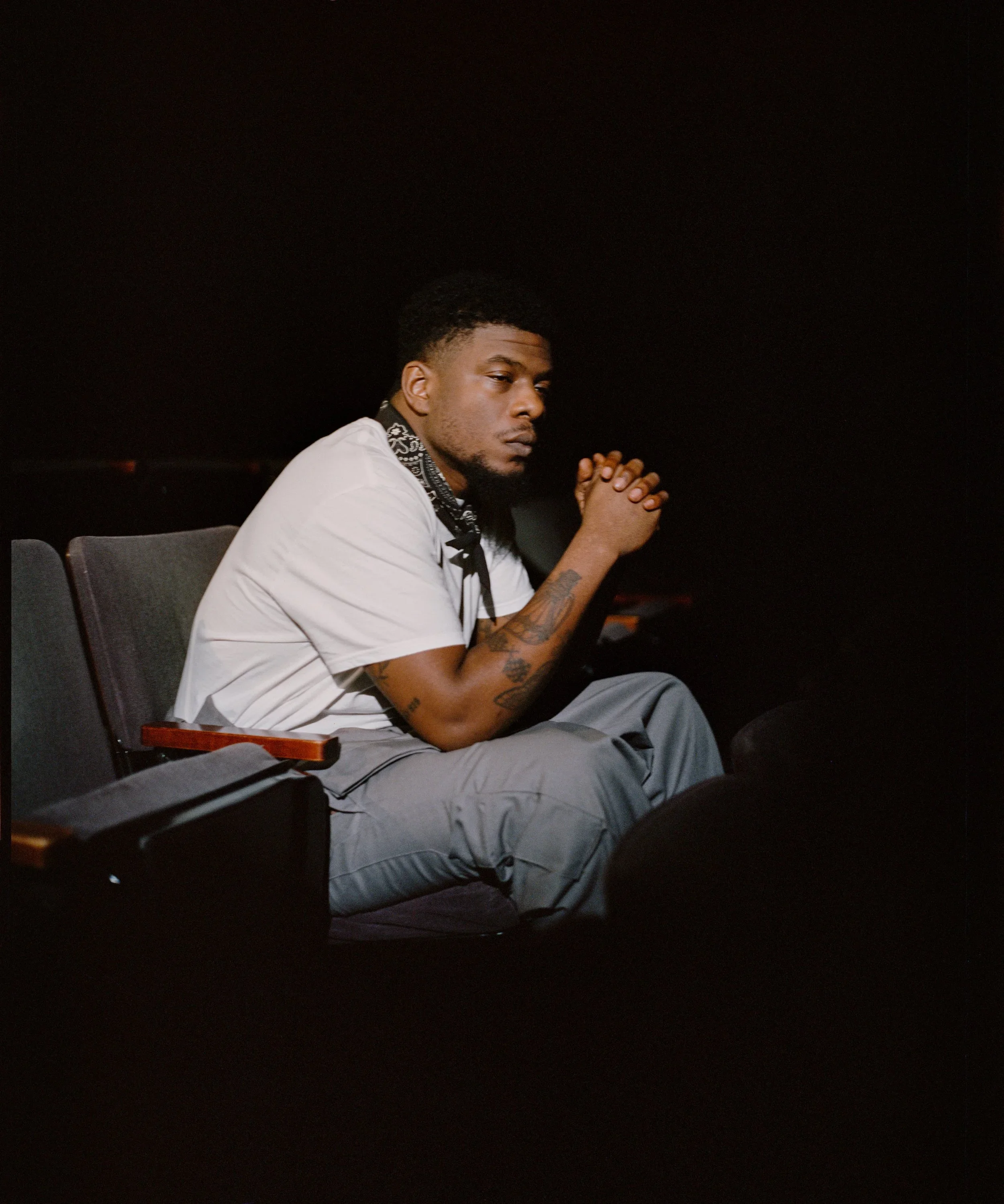Young man with tattoos sitting in a dark theater, wearing a white t-shirt and gray pants, with his hands clasped together.