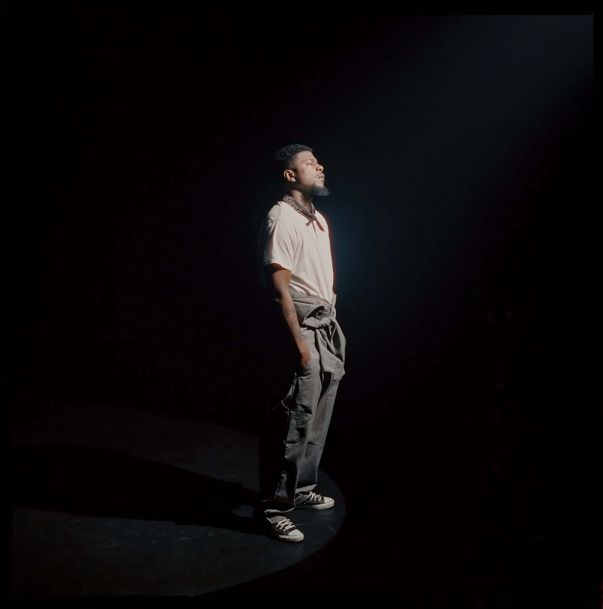 A man stands with hands in pockets in a spotlight, with a dark background.