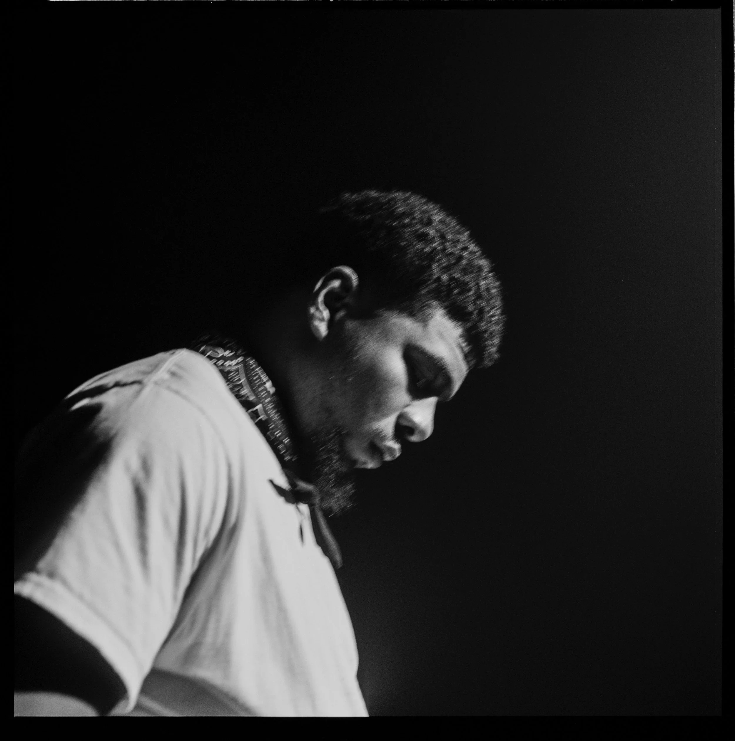 Black and white photograph of a young man in profile, with a beard and short hair, wearing a light-colored shirt and a patterned neck gaiter, looking downward against a dark background.