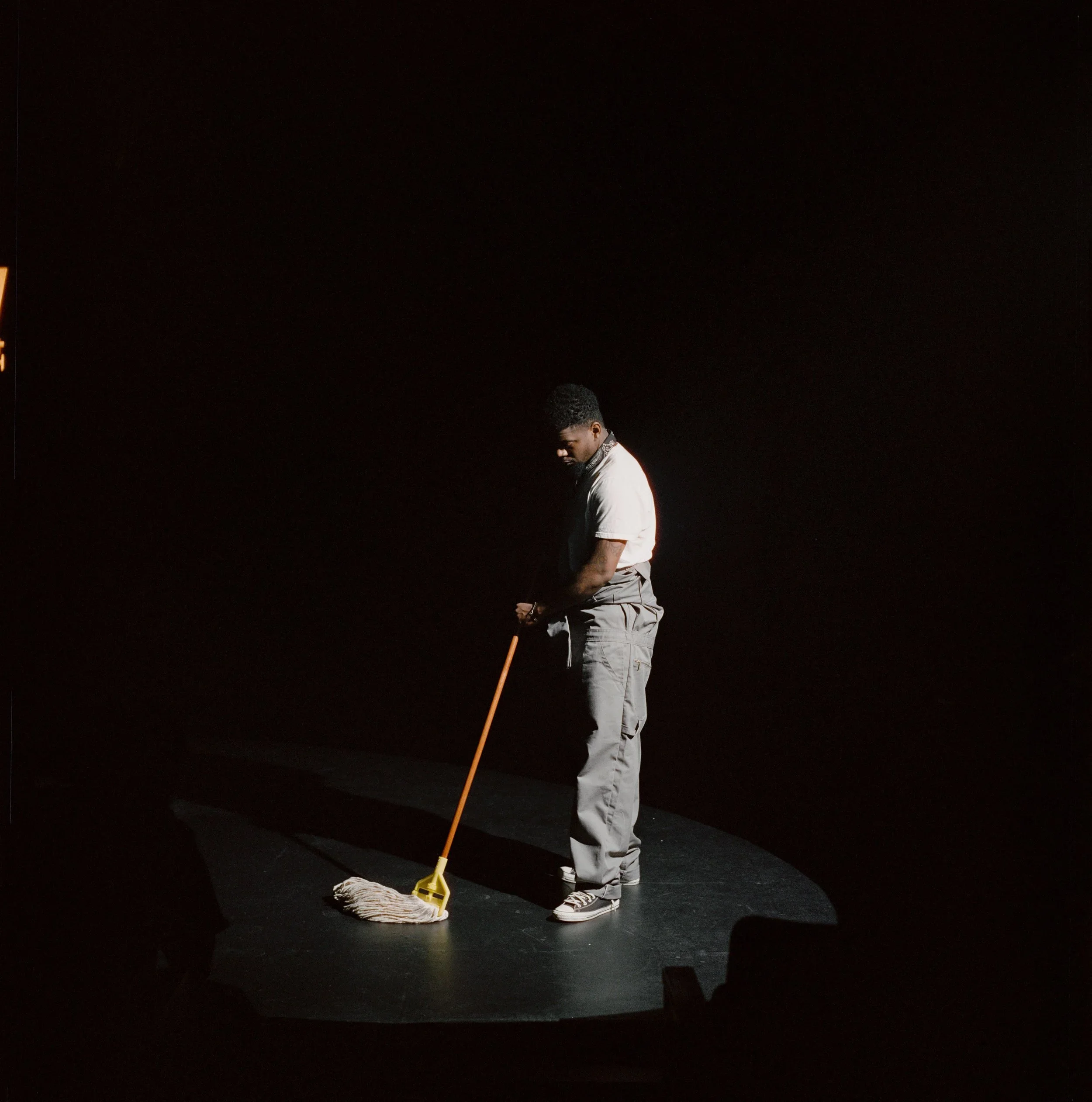 A young man with dark skin and curly hair sweeping the floor with a mop in a spotlighted area on a dark stage.