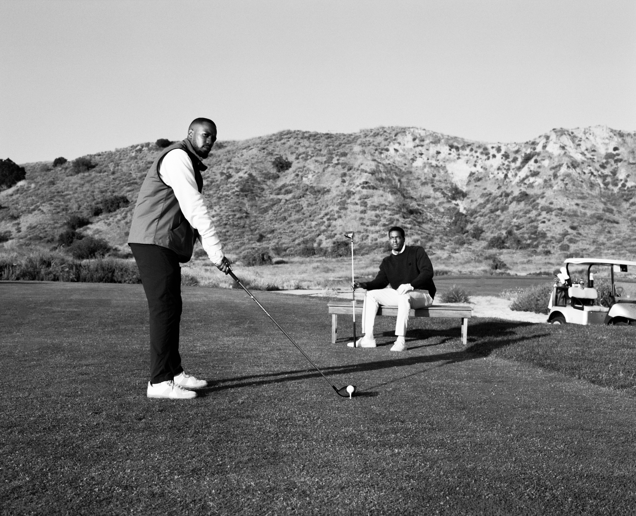 Two men at a golf course, one preparing to putt and the other sitting on a bench, with a golf cart and hilly landscape in the background.