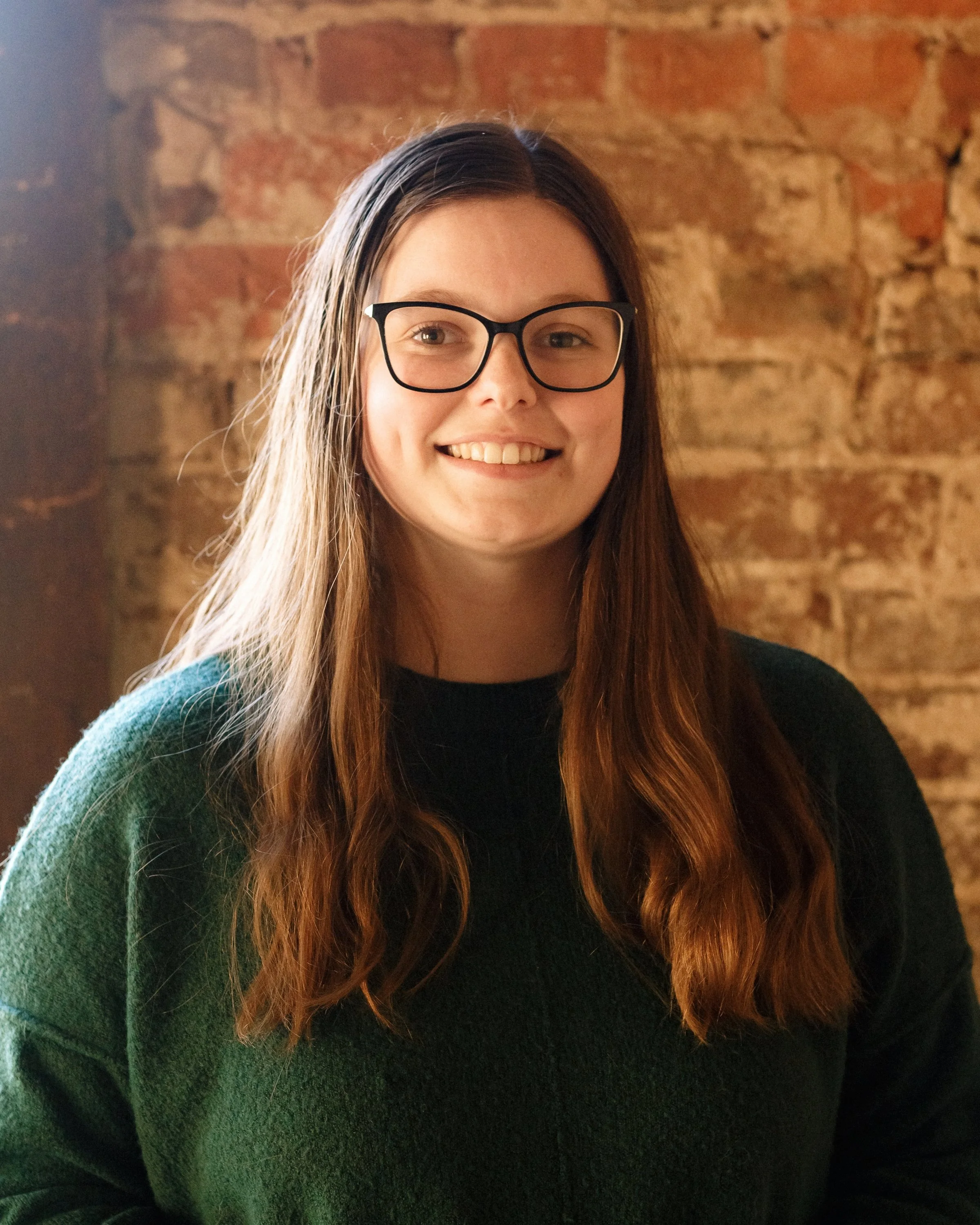 A smiling young woman with long brown hair, wearing glasses and a dark green sweater, standing in front of a brick wall.