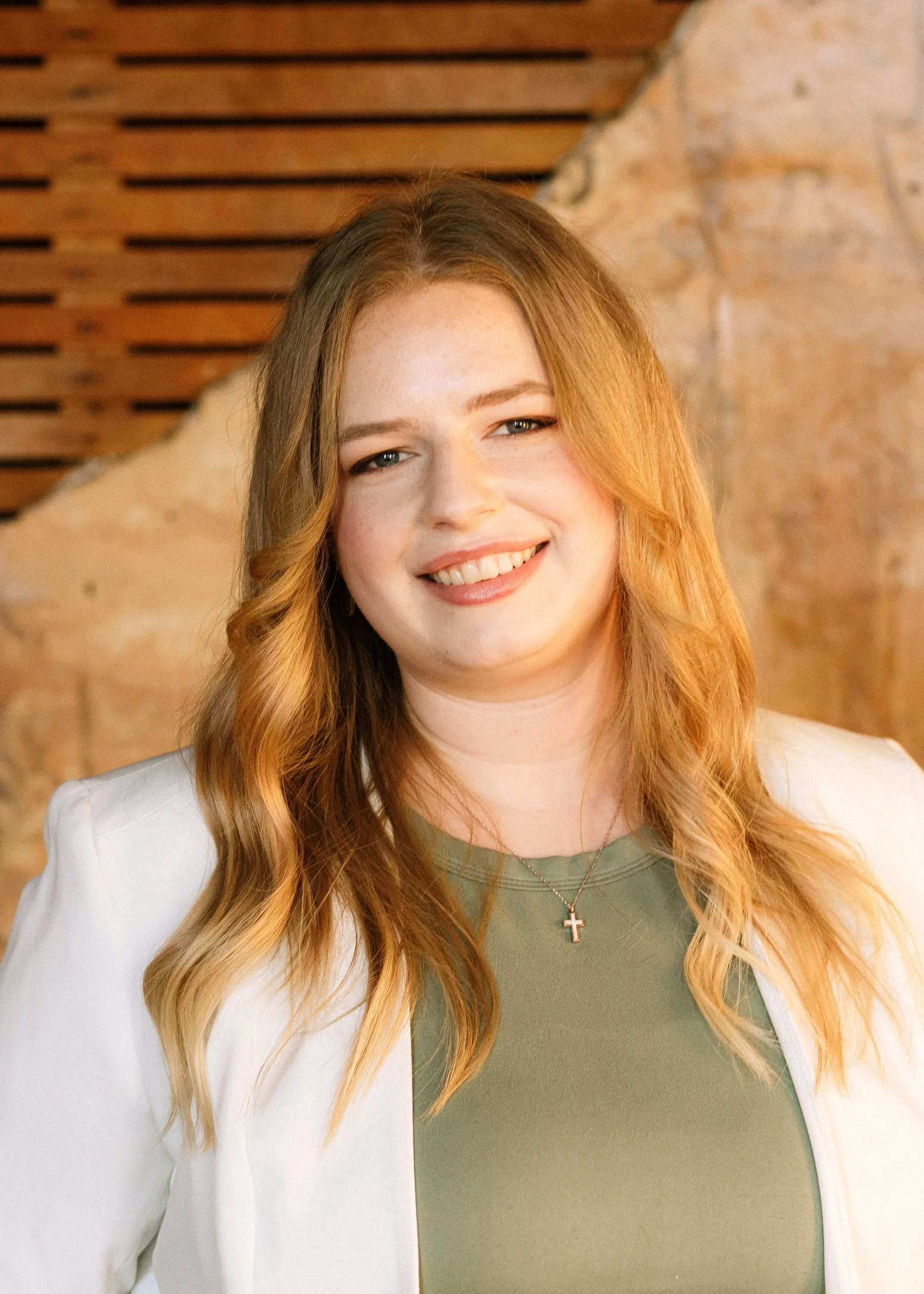 A woman with long, wavy red hair, smiling at the camera, wearing a white blazer, green top, a cross necklace, standing in front of a stone and wood background.