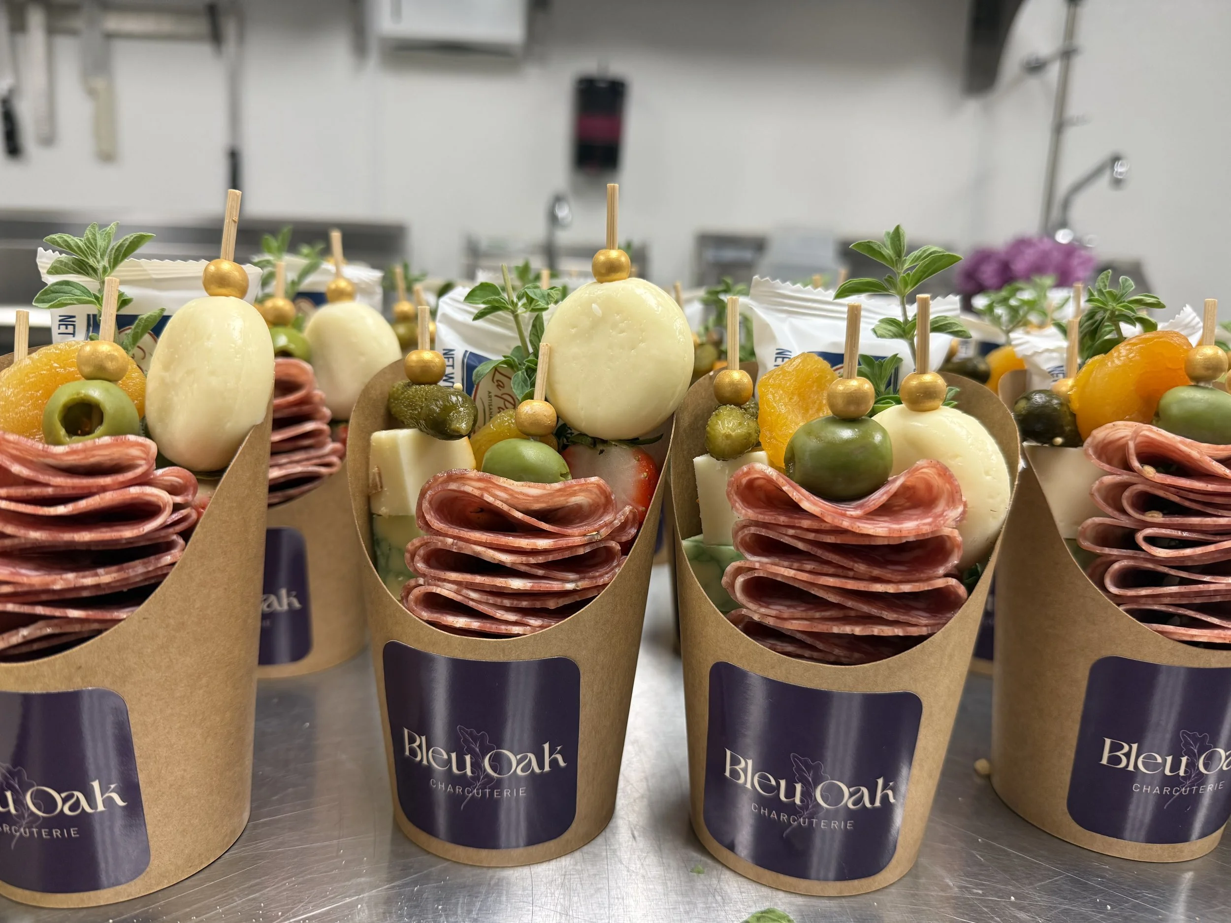 Charcuterie cups with stacked salami slices, cheese, olives, pickles, fruit, and greenery, labeled 'Bleu Oak'.