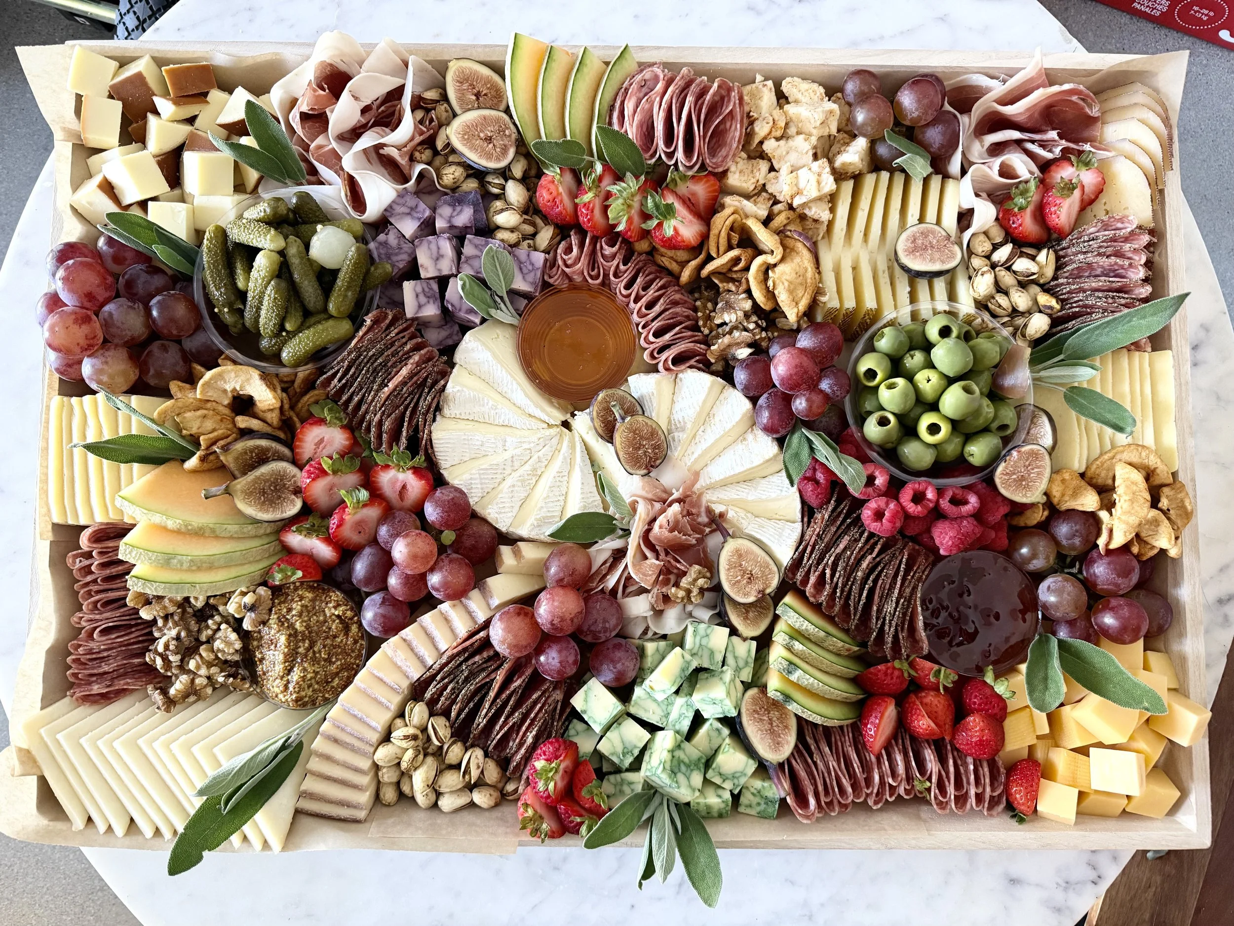 A large wooden charcuterie board with assorted cheeses, cured meats, fruits, nuts, and garnishes arranged in sections.
