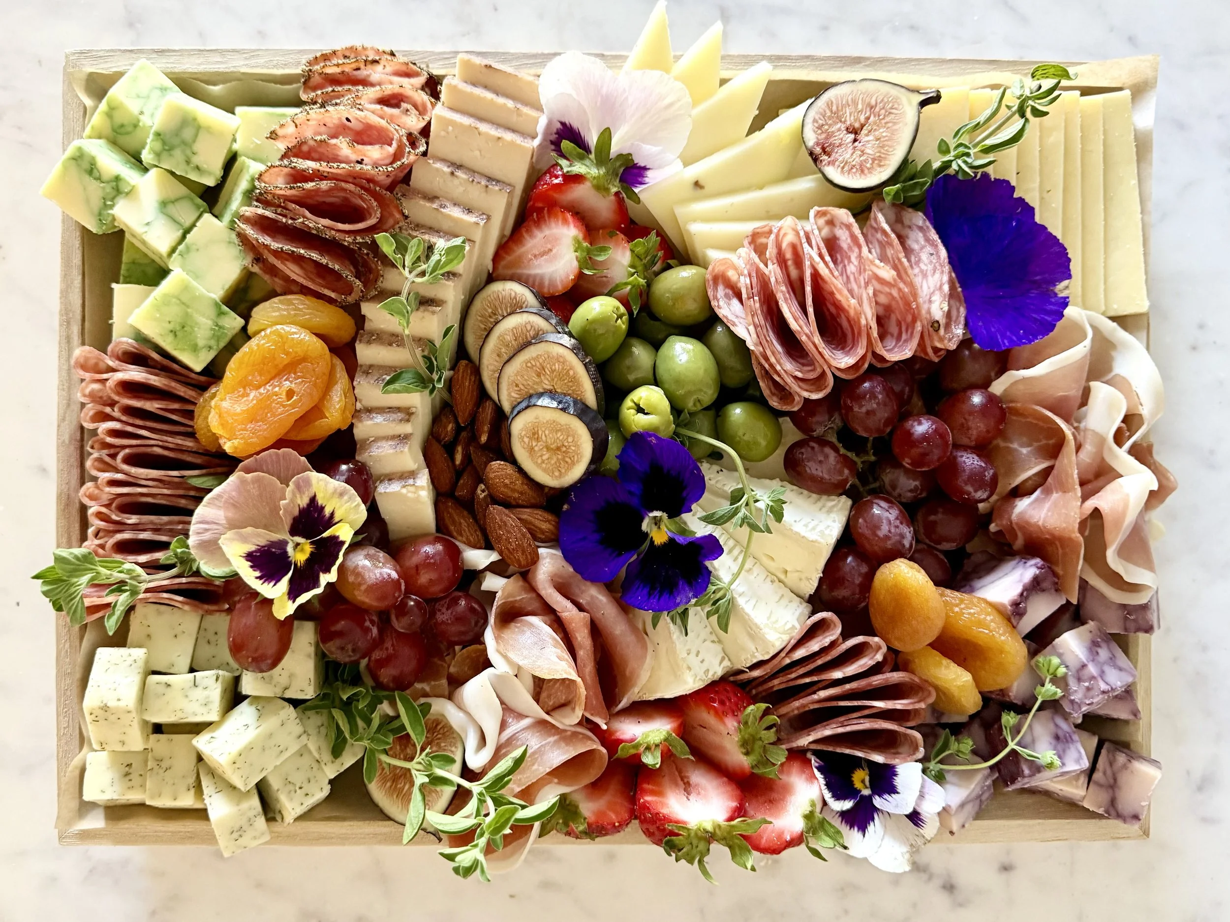 A wooden cheese and charcuterie board with assorted cheeses, cured meats, fresh strawberries, grapes, figs, almonds, dried apricots, garnished with edible flowers and herbs.