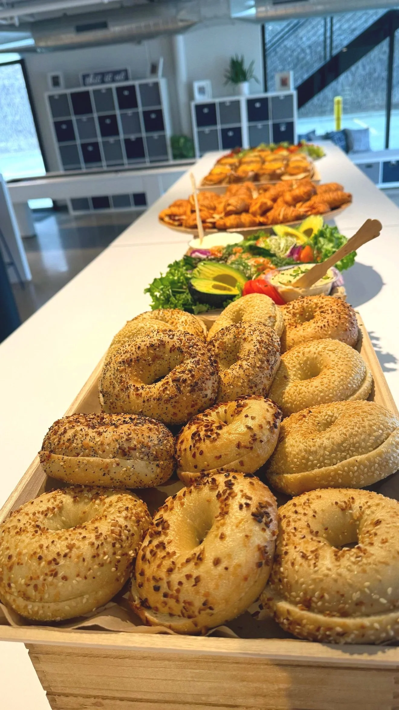 Fresh bagels topped with various seeds and spices arranged on a wooden tray at a buffet table.