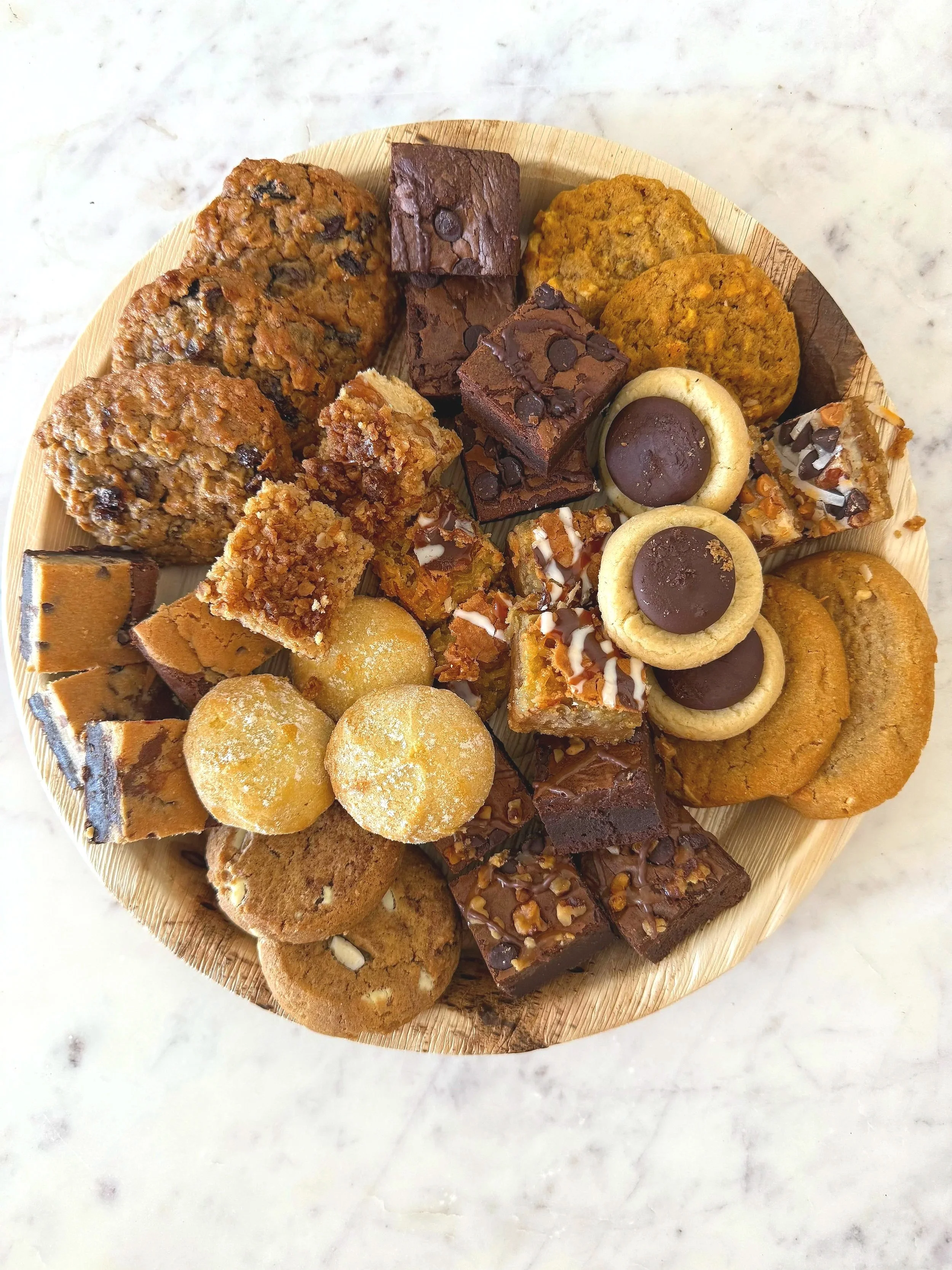 A round wooden platter filled with assorted cookies, brownies, and sweet treats on a white marble surface.