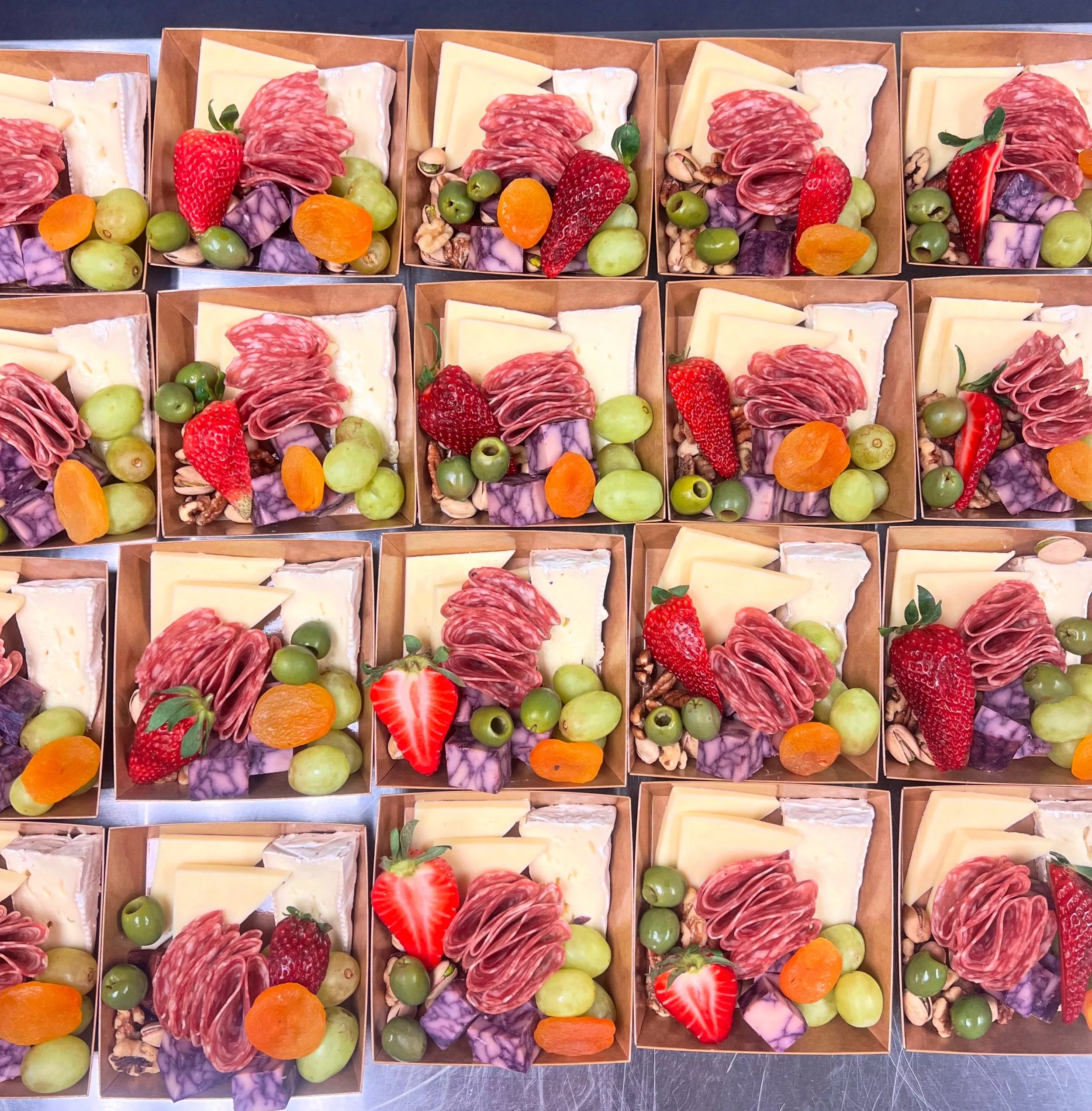 Assorted charcuterie boxes with cheese, cured meat, strawberries, grapes, dried apricots, nuts, and purple cabbage, arranged in a grid.