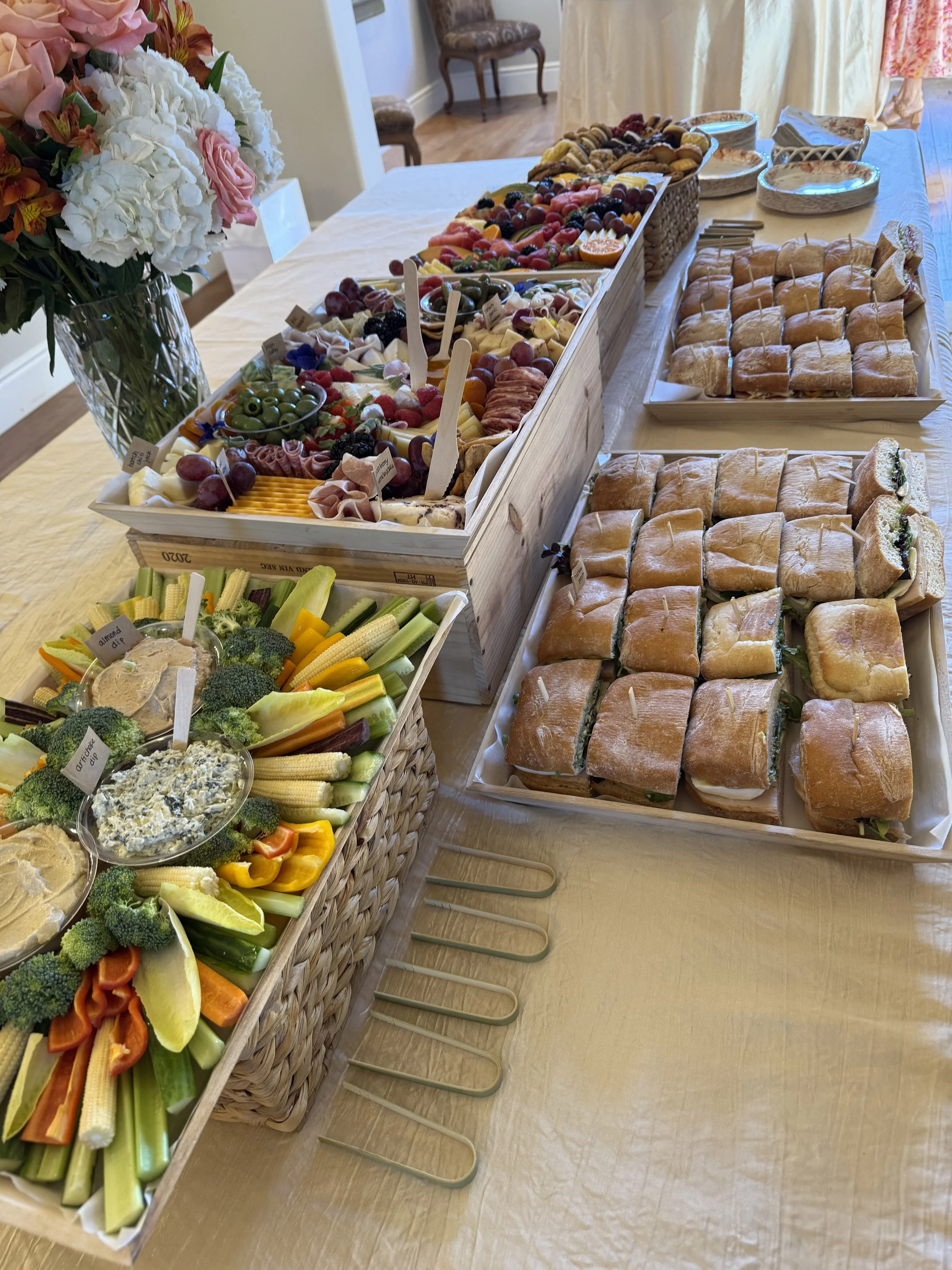 A buffet table with an assortment of food items, including a large tray of mini sandwiches, vegetable platter with dips, a fruit and cheese platter, and a bouquet of flowers in a glass vase.