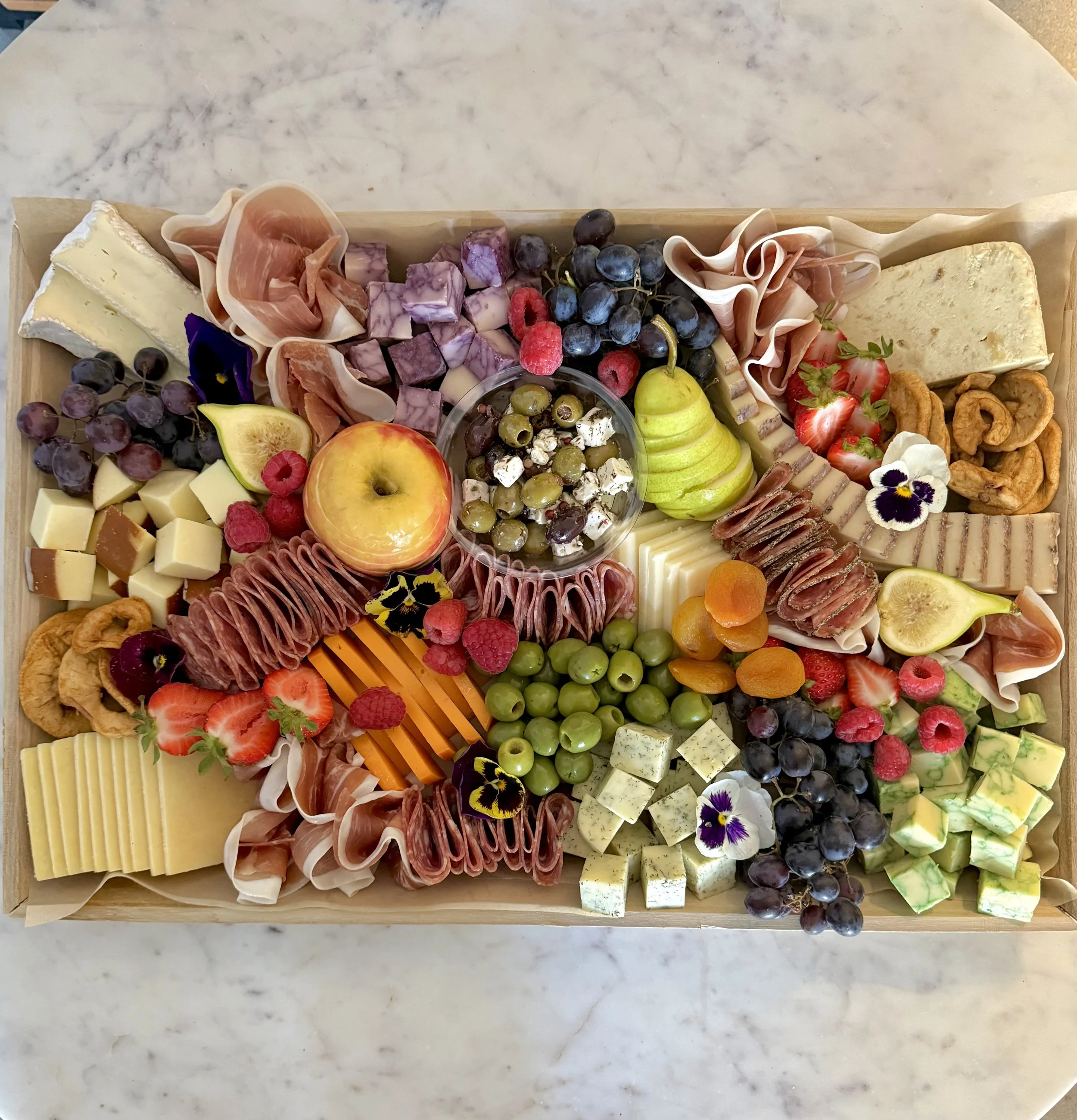 A cheese and charcuterie board with grapes, strawberries, figs, raspberries, peaches, and apricots, accompanied by assorted cheeses, cured meats, nuts, and edible flowers.