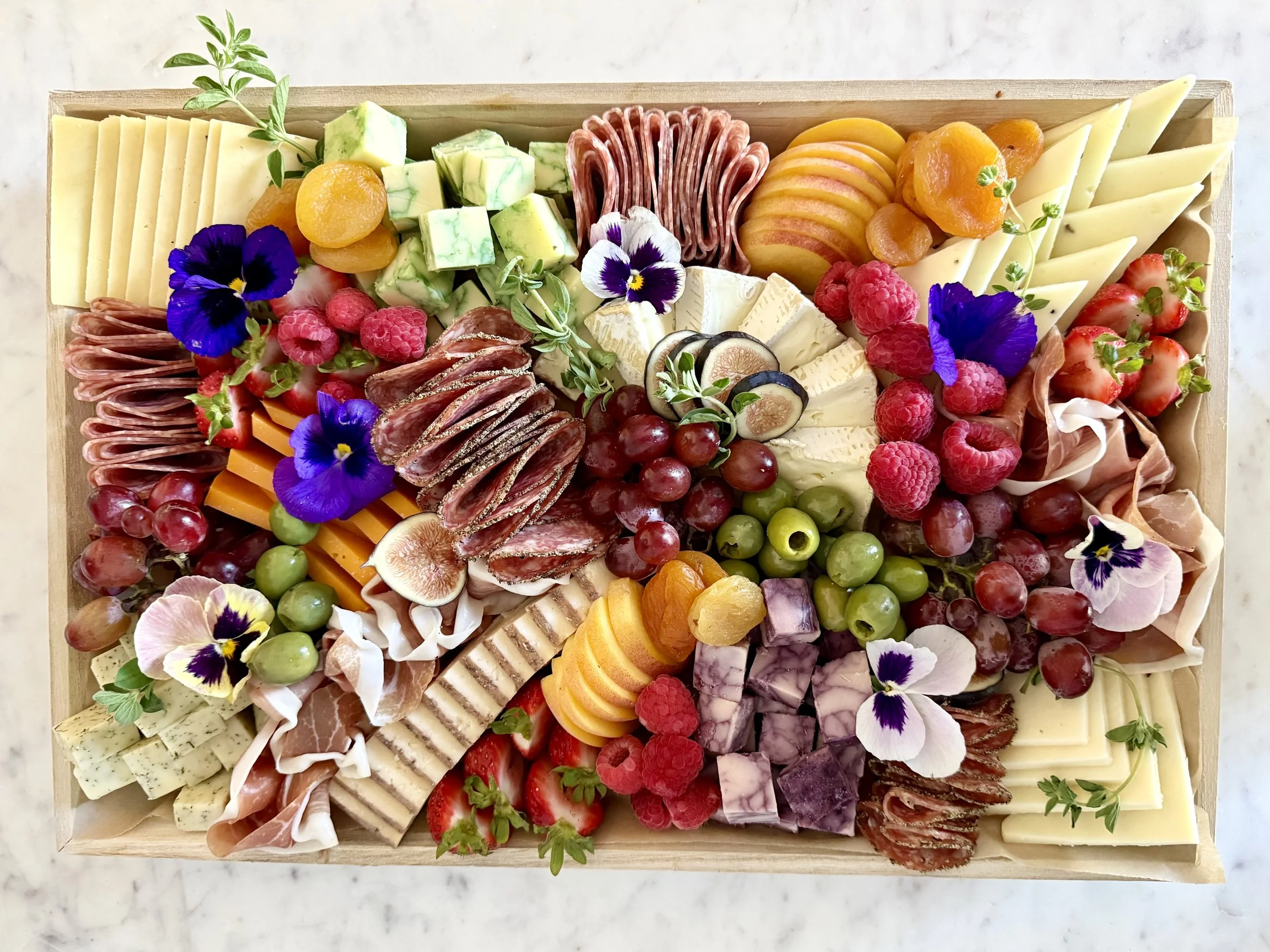 A wooden cheese and charcuterie platter with various cheeses, cured meats, grapes, strawberries, raspberries, figs, apricots, orange slices, and edible flowers.