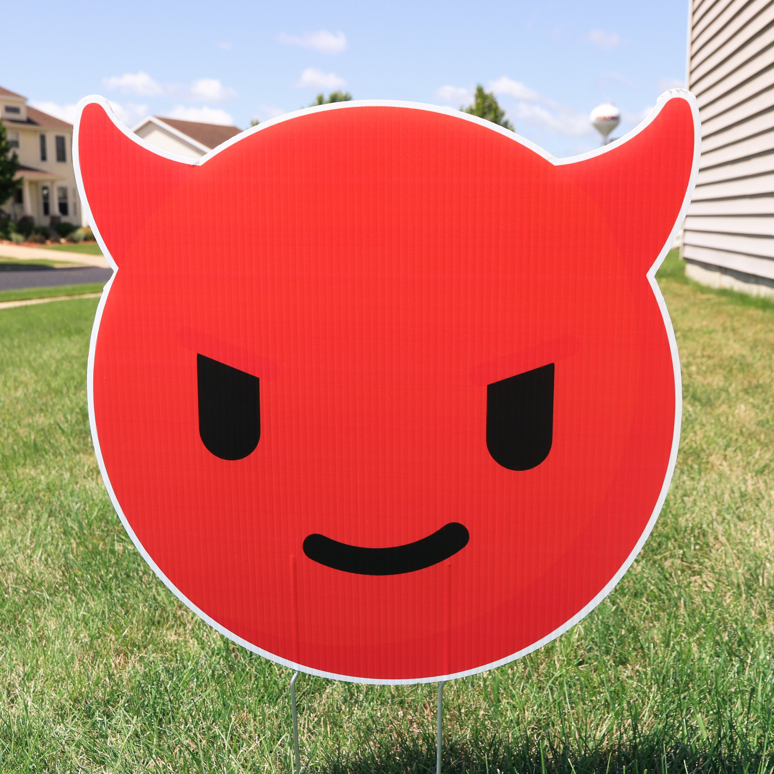 Red devil emoji yard sign with horns in a grassy area near houses.