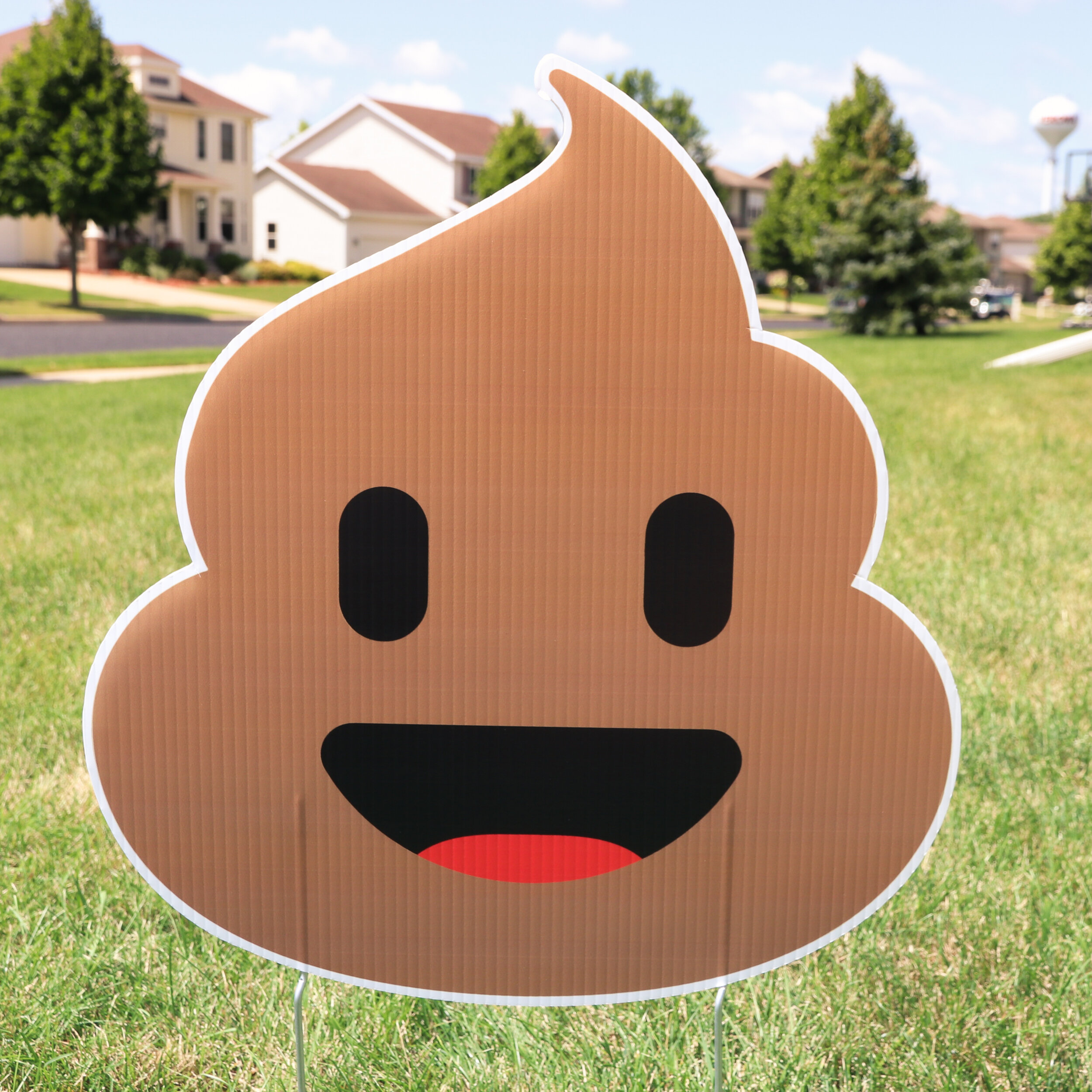 A cardboard cutout of a smiling poop emoji with eyes and a mouth, placed on a grassy lawn in a suburban neighborhood.