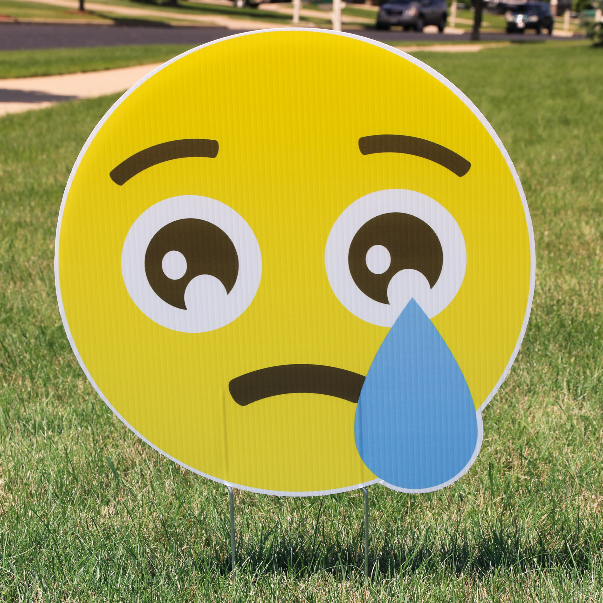 A large yellow emoji lawn sign displaying a sad face with a single tear, placed on green grass.