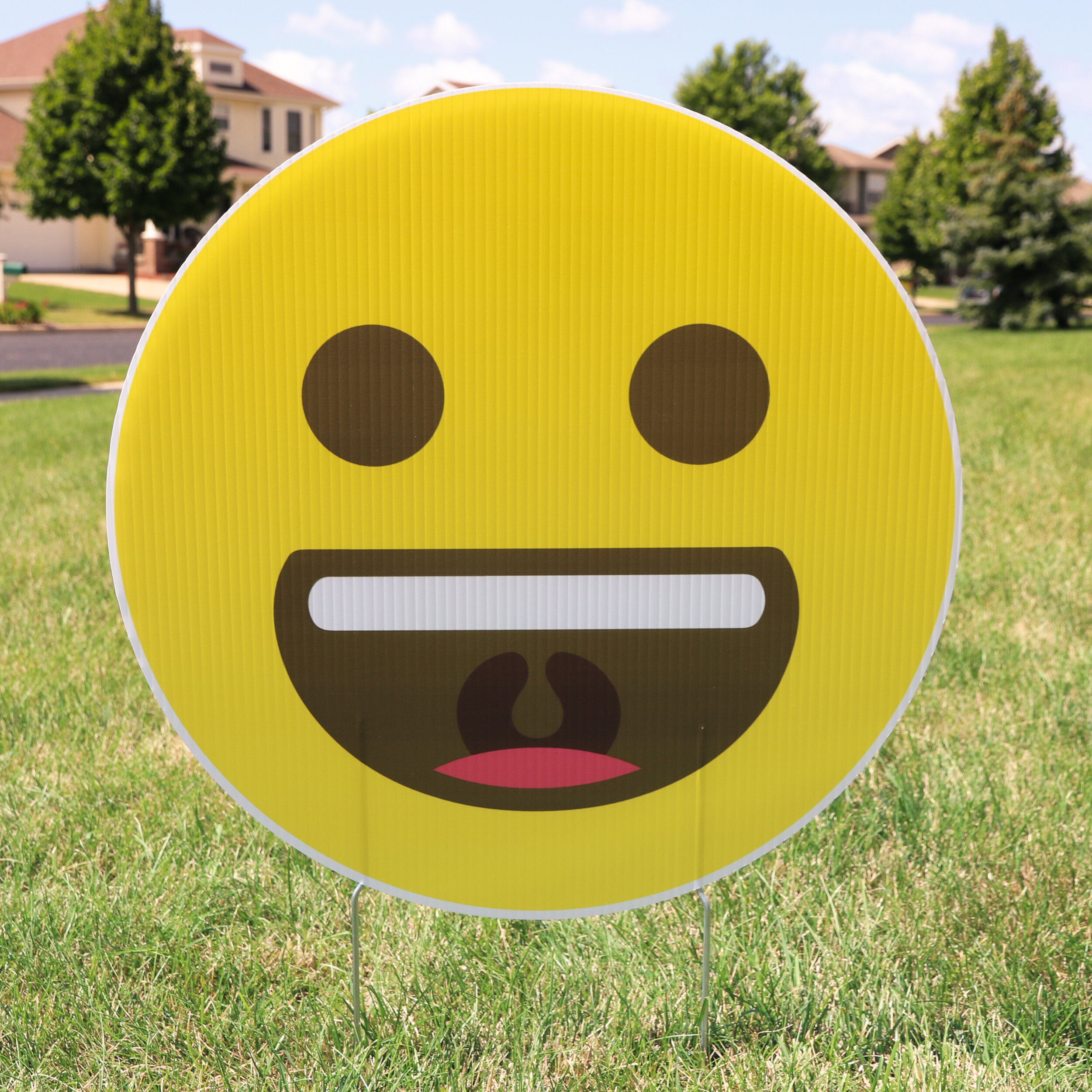 Large smiling emoji yard sign on green grass with houses and trees in the background.