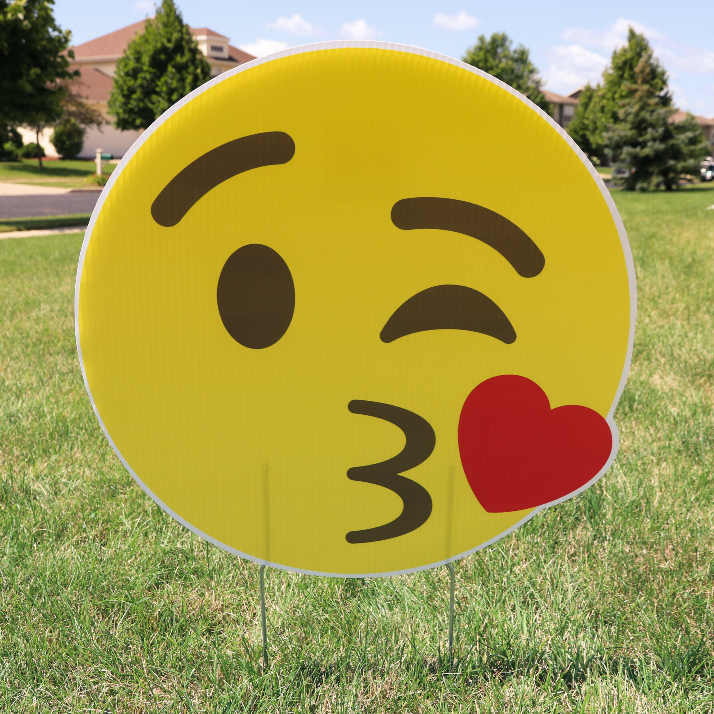 Large emoji face sign with winking kiss and heart on grass, background with trees and houses.