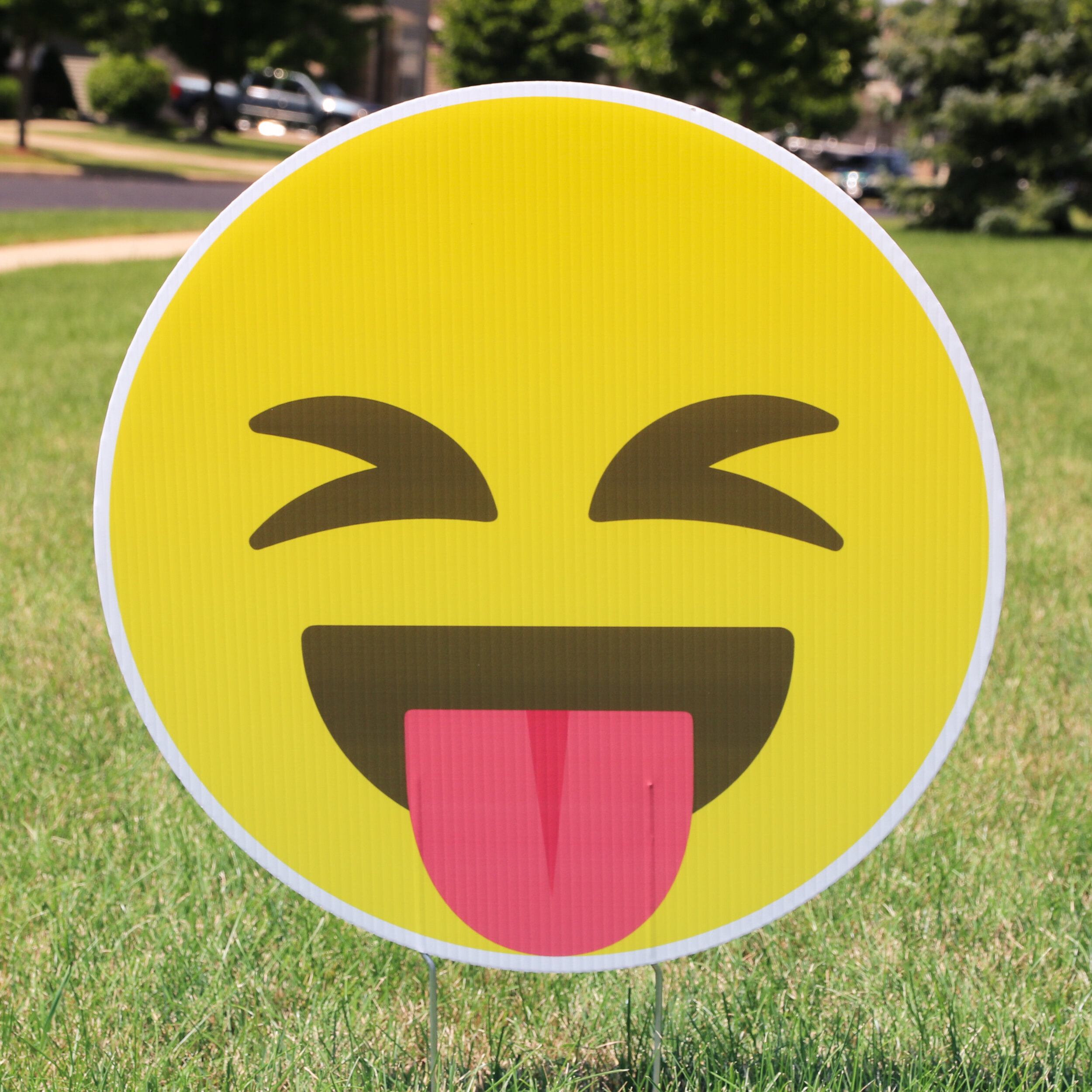 Large yellow emoji sign with closed eyes and sticking out tongue, placed on a grassy lawn.