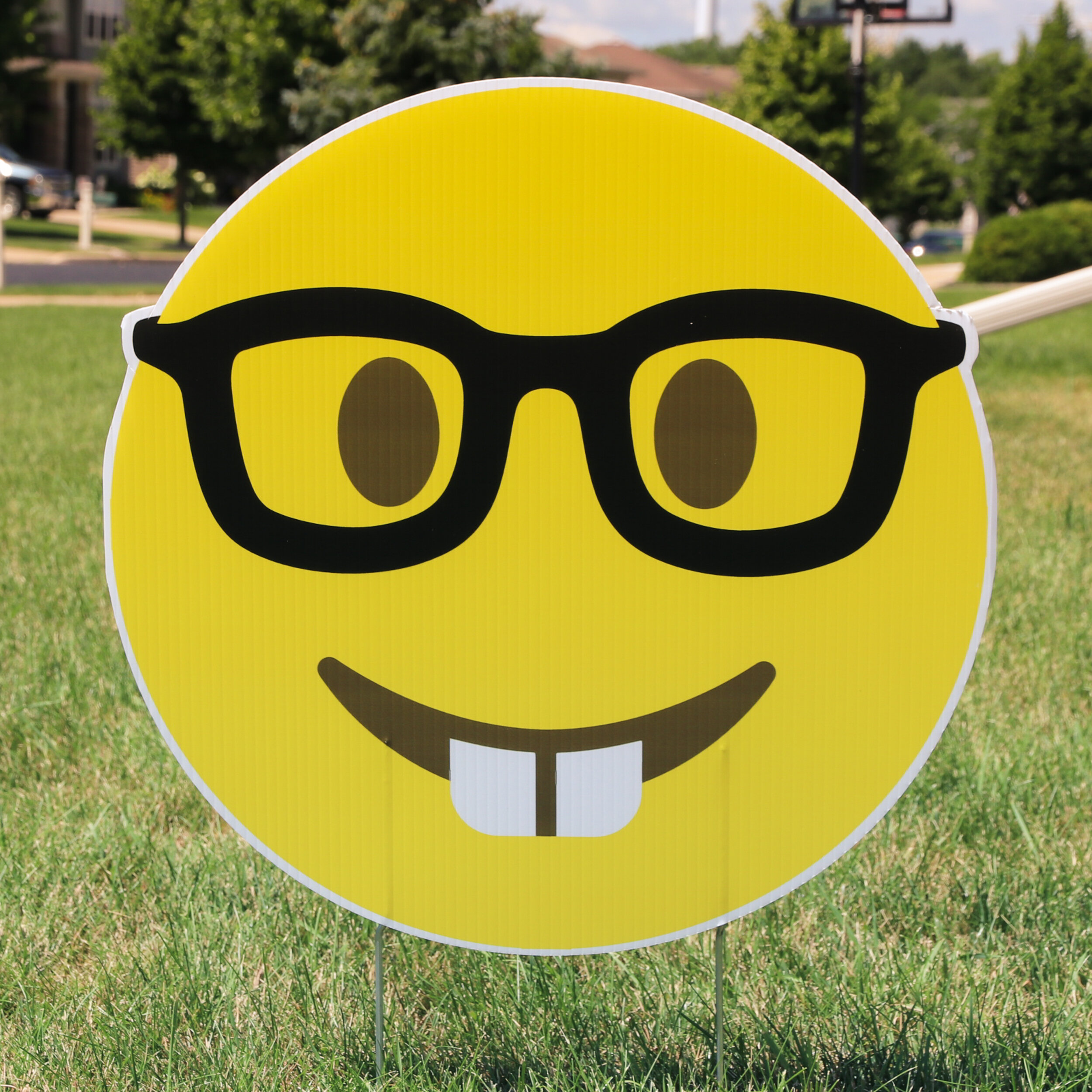 A yellow smiley face sign with black glasses and protruding teeth on a grassy lawn.
