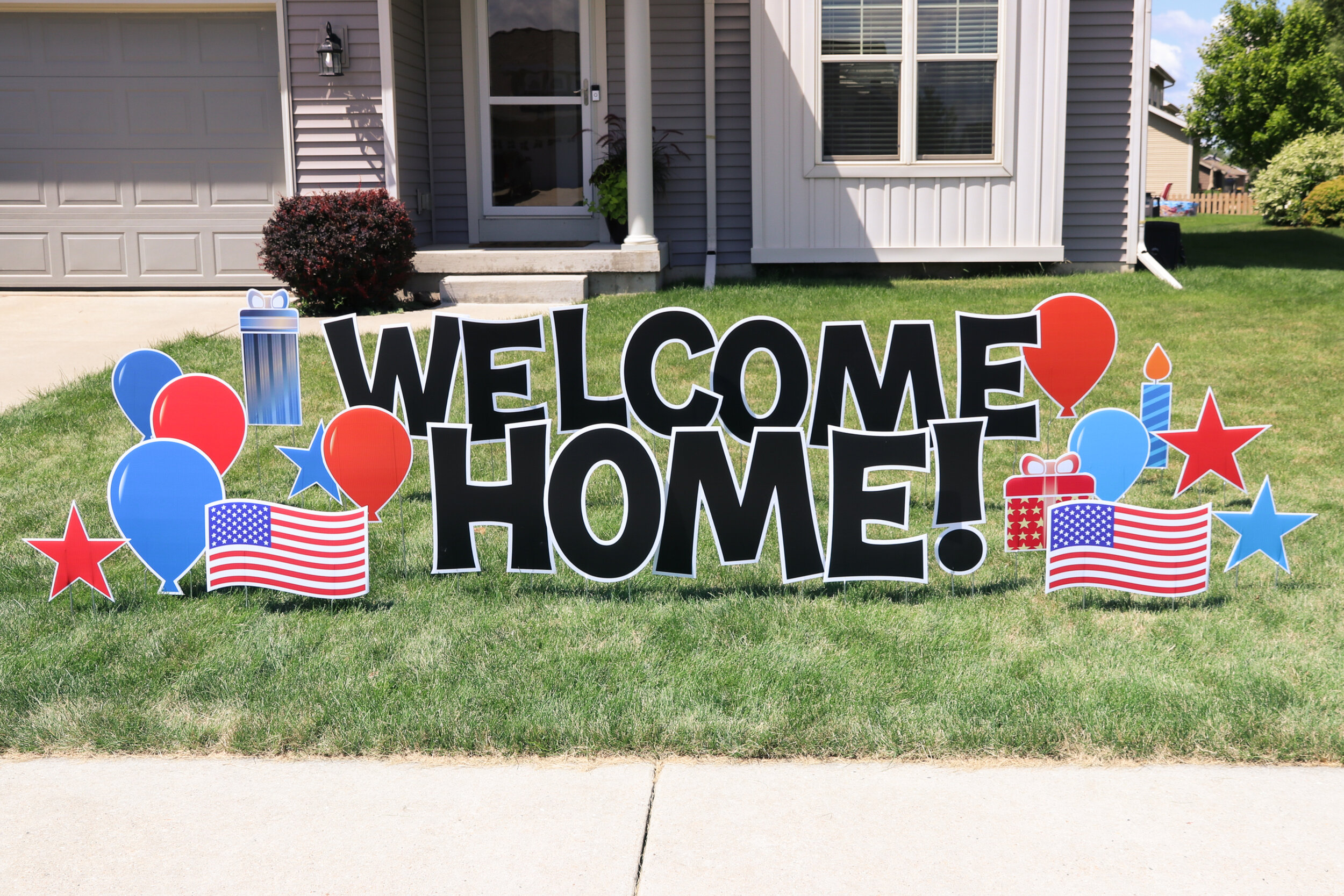 "Welcome Home" sign with red, white, and blue balloons, stars, American flags, and gift icons on a lawn in front of a house.