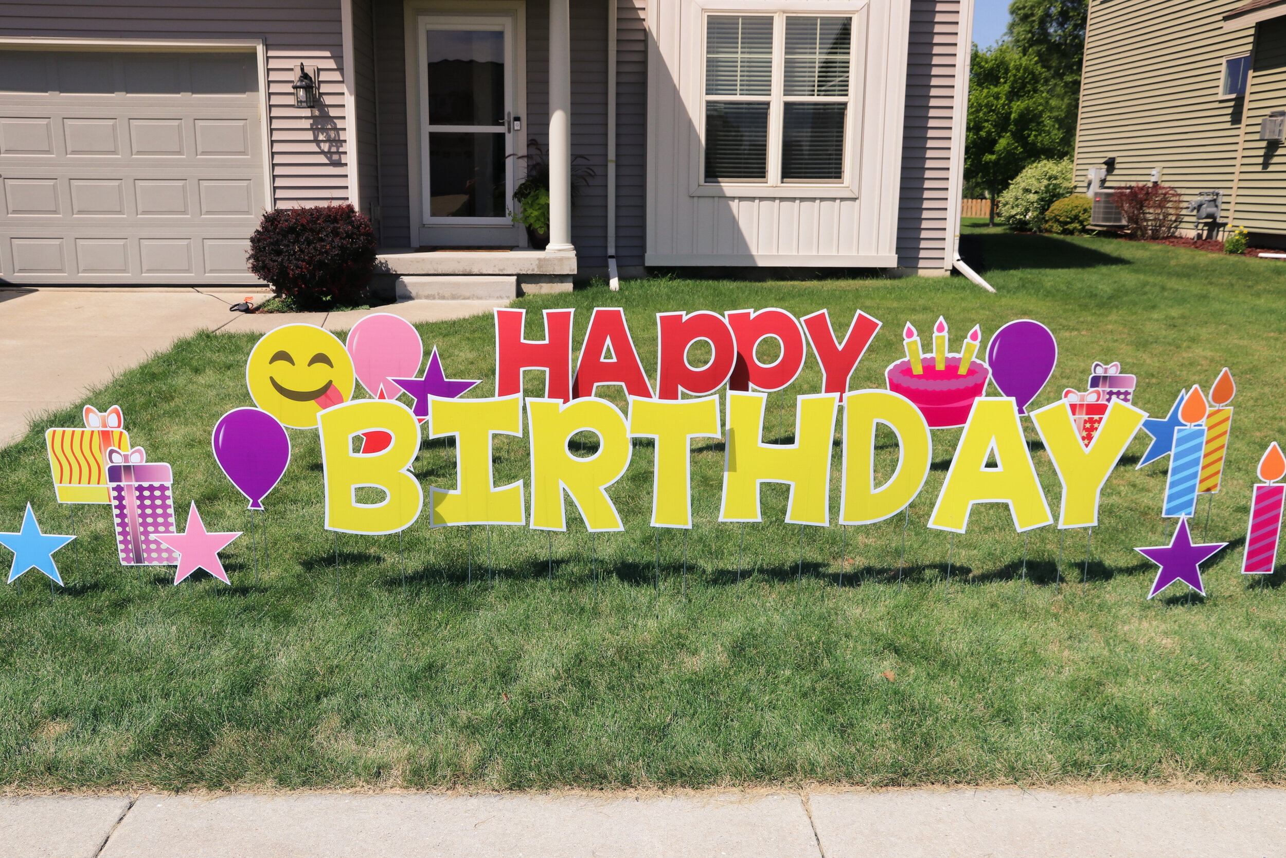 Lawn display with colorful 'Happy Birthday' sign featuring balloons, stars, presents, and a cake in front of a house.