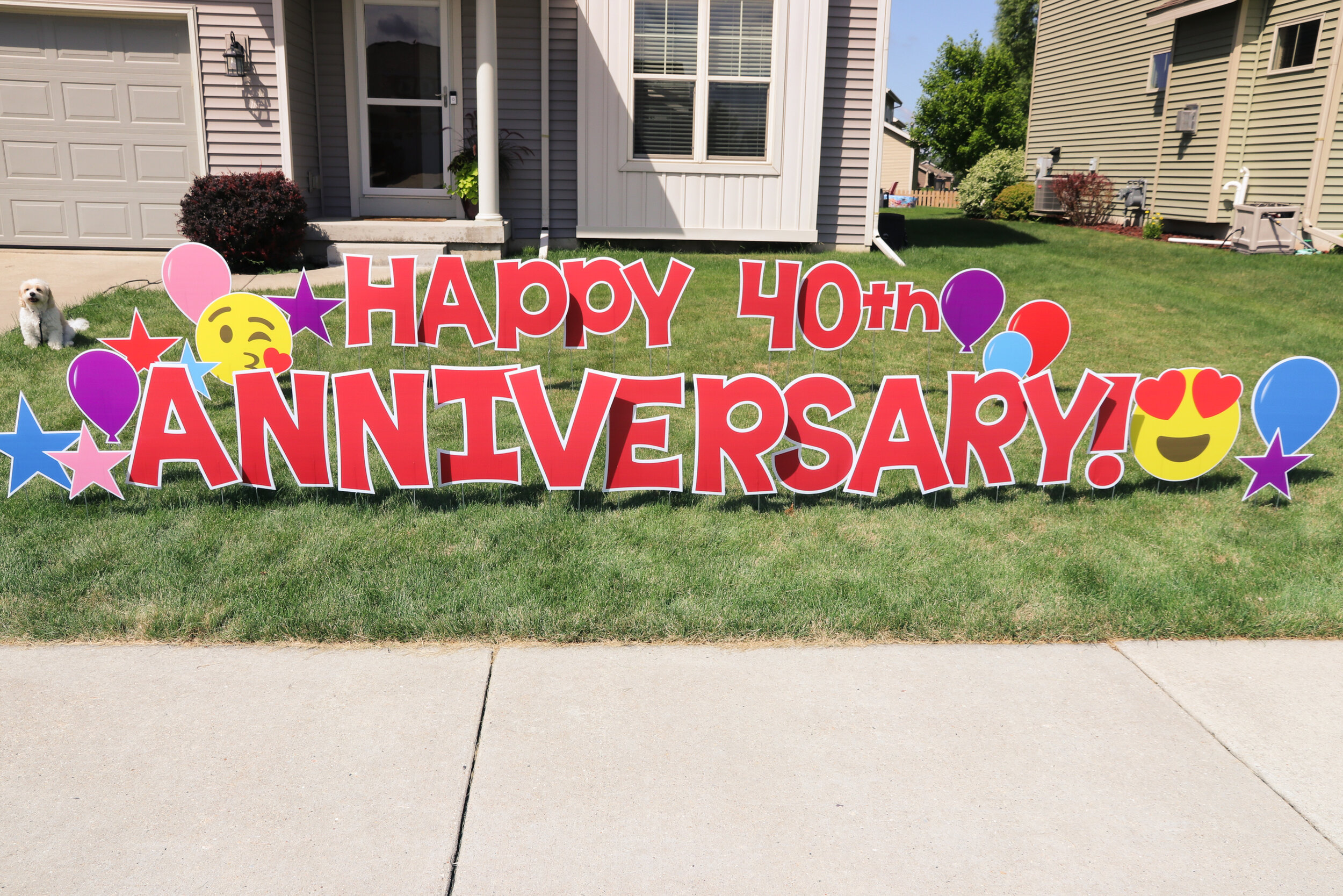Outdoor yard sign reading 'Happy 40th Anniversary' with balloons and emojis, in front of a house.