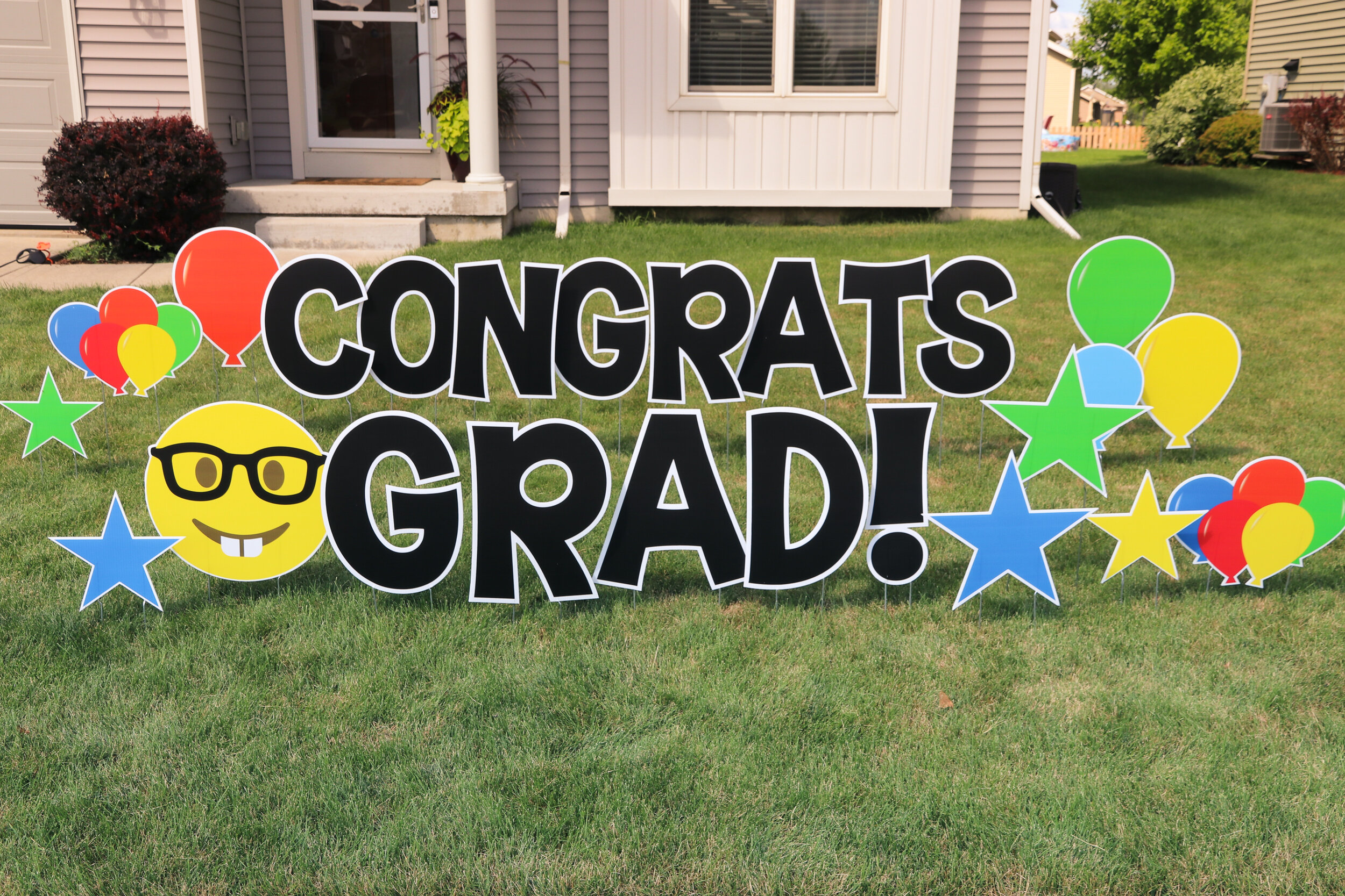 Lawn sign with "Congrats Grad!" message, colorful balloons, stars, and smiley face with glasses.