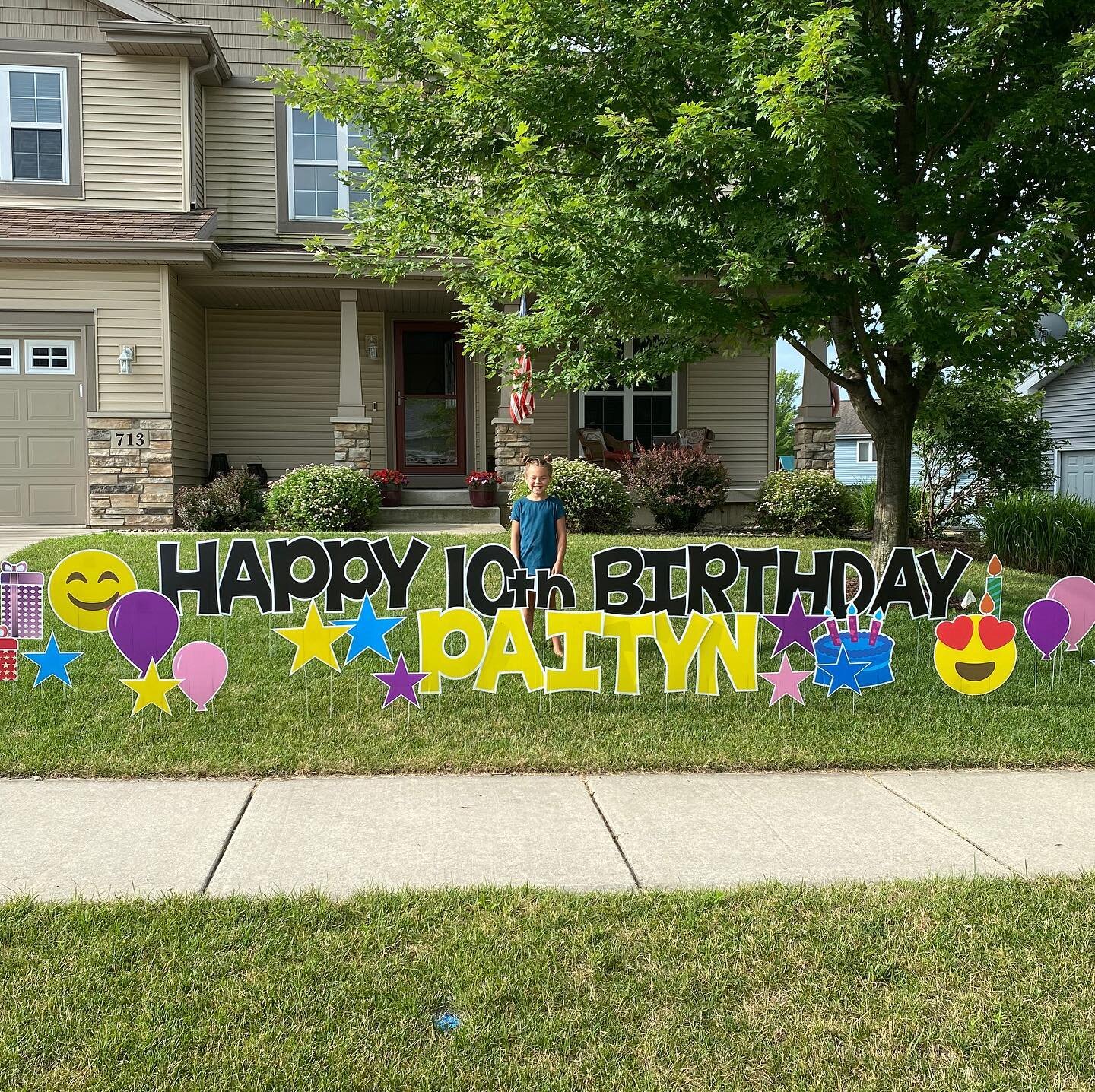 A child standing in front of a house with a lawn sign reading "Happy 10th Birthday Paityn," decorated with balloons, stars, and emojis.