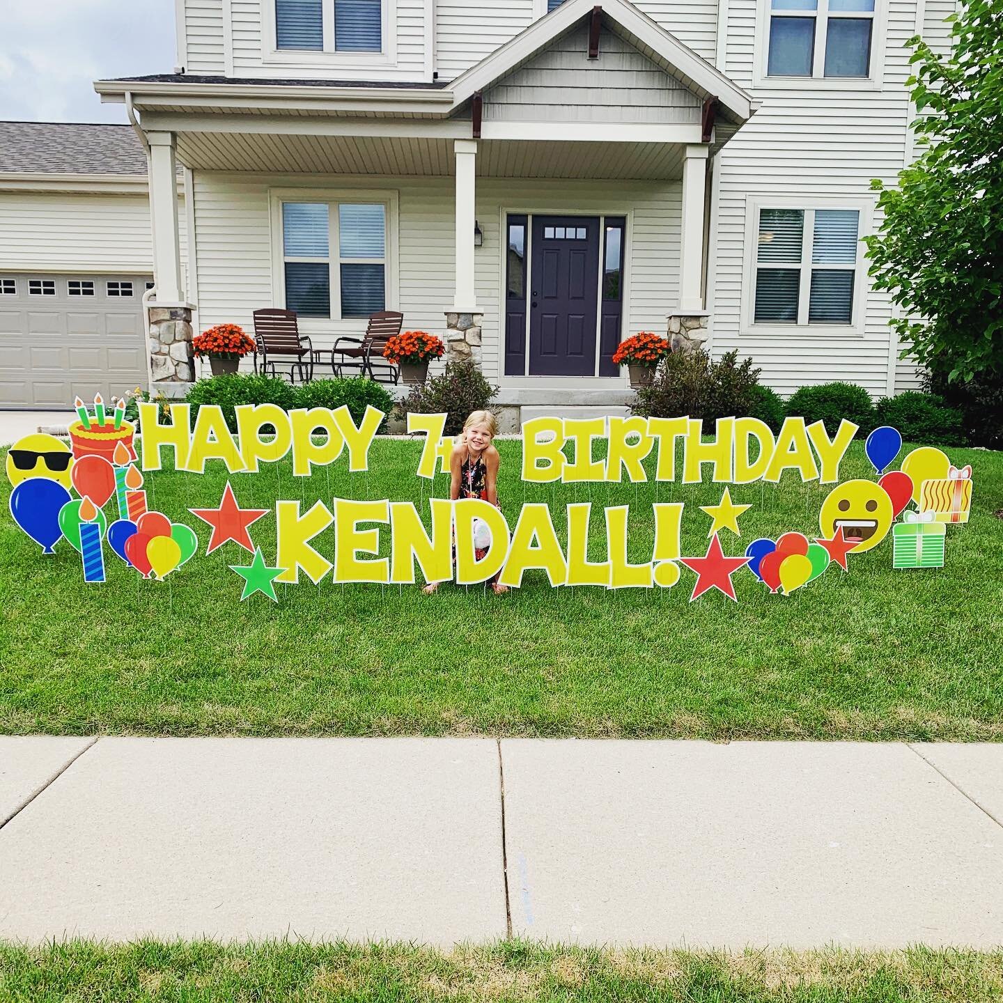 Large colorful yard sign in front of a house with text "Happy 7th Birthday Kendall!" and a child crouching next to it.