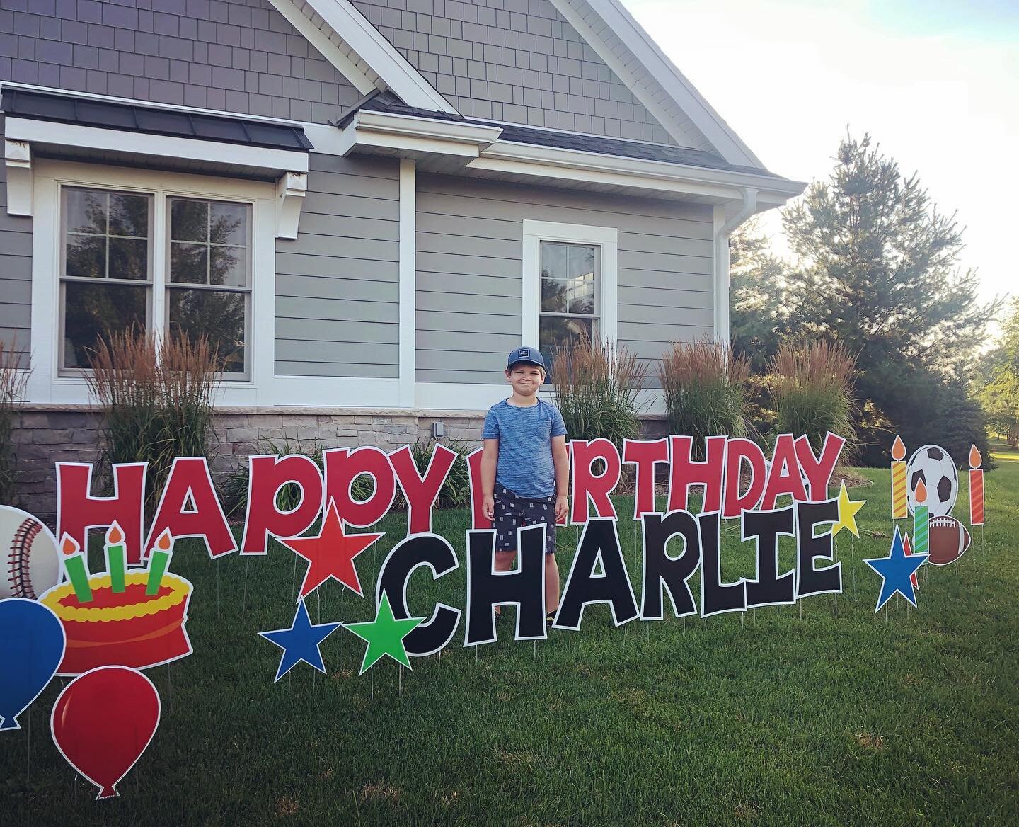 Child standing in front of a house with large yard signs spelling "Happy Birthday Charlie," featuring balloons, stars, cupcakes, and sports balls.