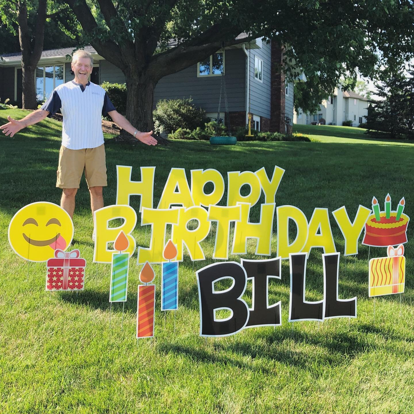Man standing in front of a large outdoor sign reading 'Happy Birthday Bill' with festive emojis, candles, and gifts, on a grassy lawn with trees and a house in the background.