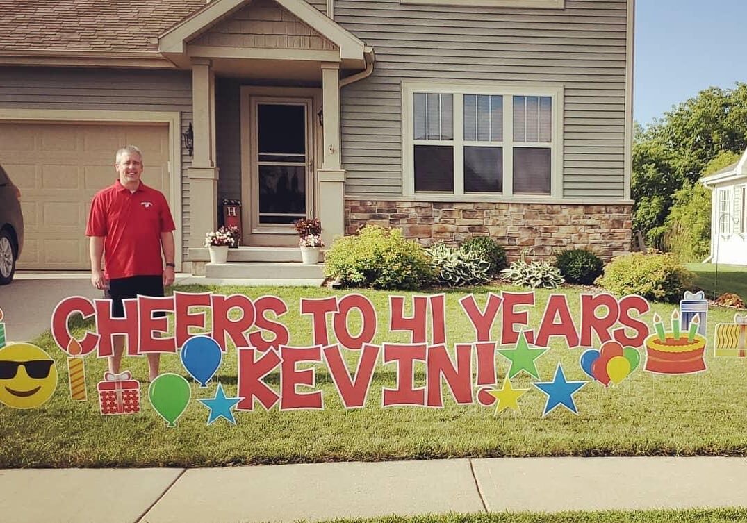 Man in red shirt standing by colorful yard sign reading "Cheers to 41 Years Kevin" with balloons and cake graphics in front of a house.