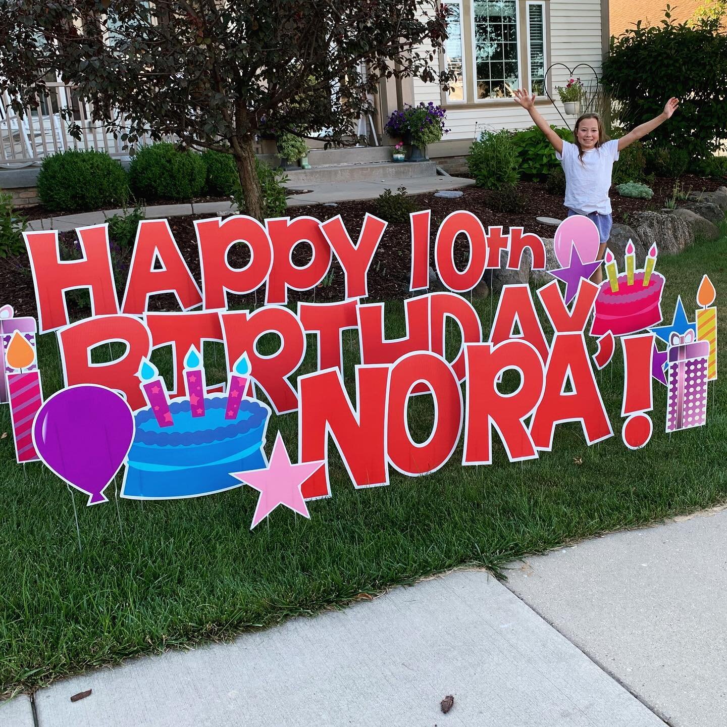 Yard sign displaying 'Happy 10th Birthday Nora' with a child standing nearby. Includes birthday-themed decorations like cakes, candles, and balloons, set in a grassy residential yard.
